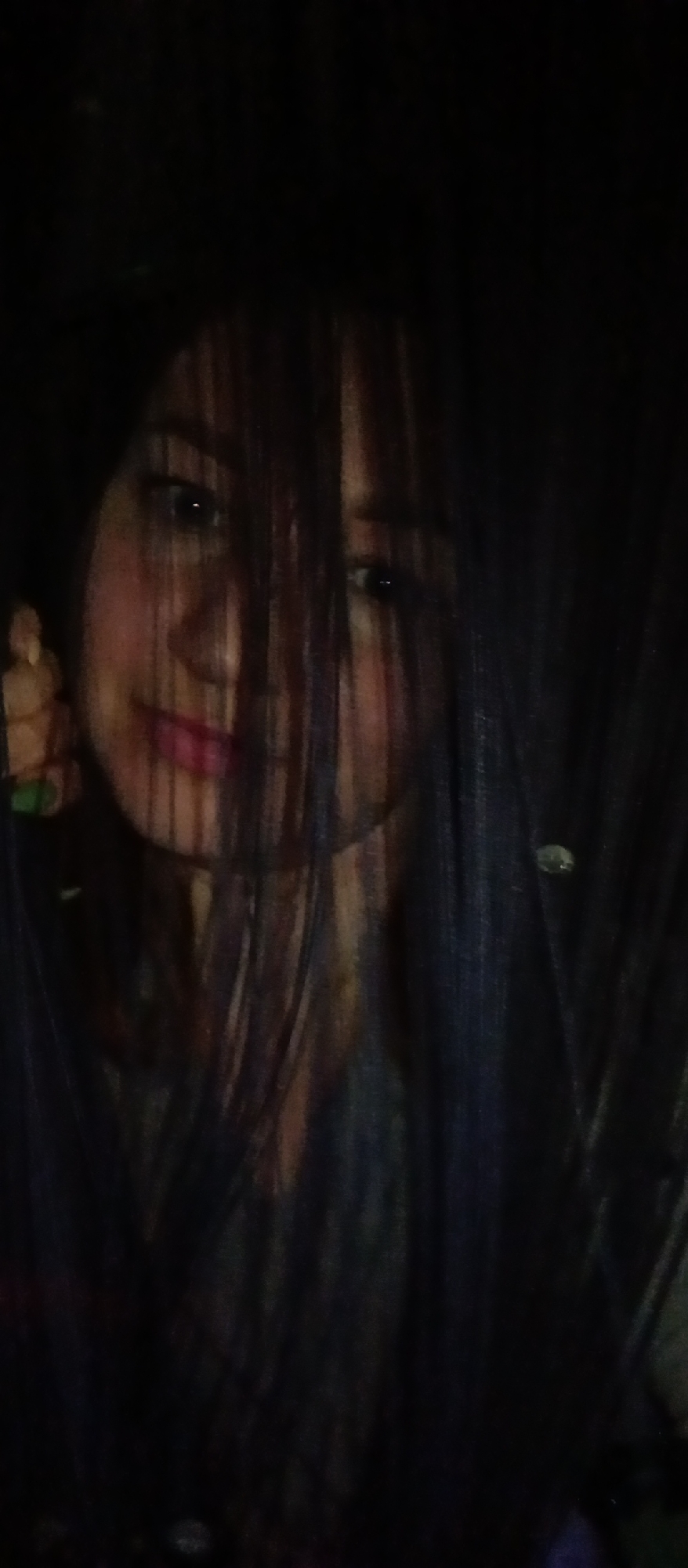 A person with dark hair obscuring their face is the primary subject of the image. The person's face is partially visible through strands of hair, revealing eyes, nose, and lips. The background is dark, with a few indistinct objects. It is difficult to determine a location or identify specific objects due to the low lighting and obstructed view.