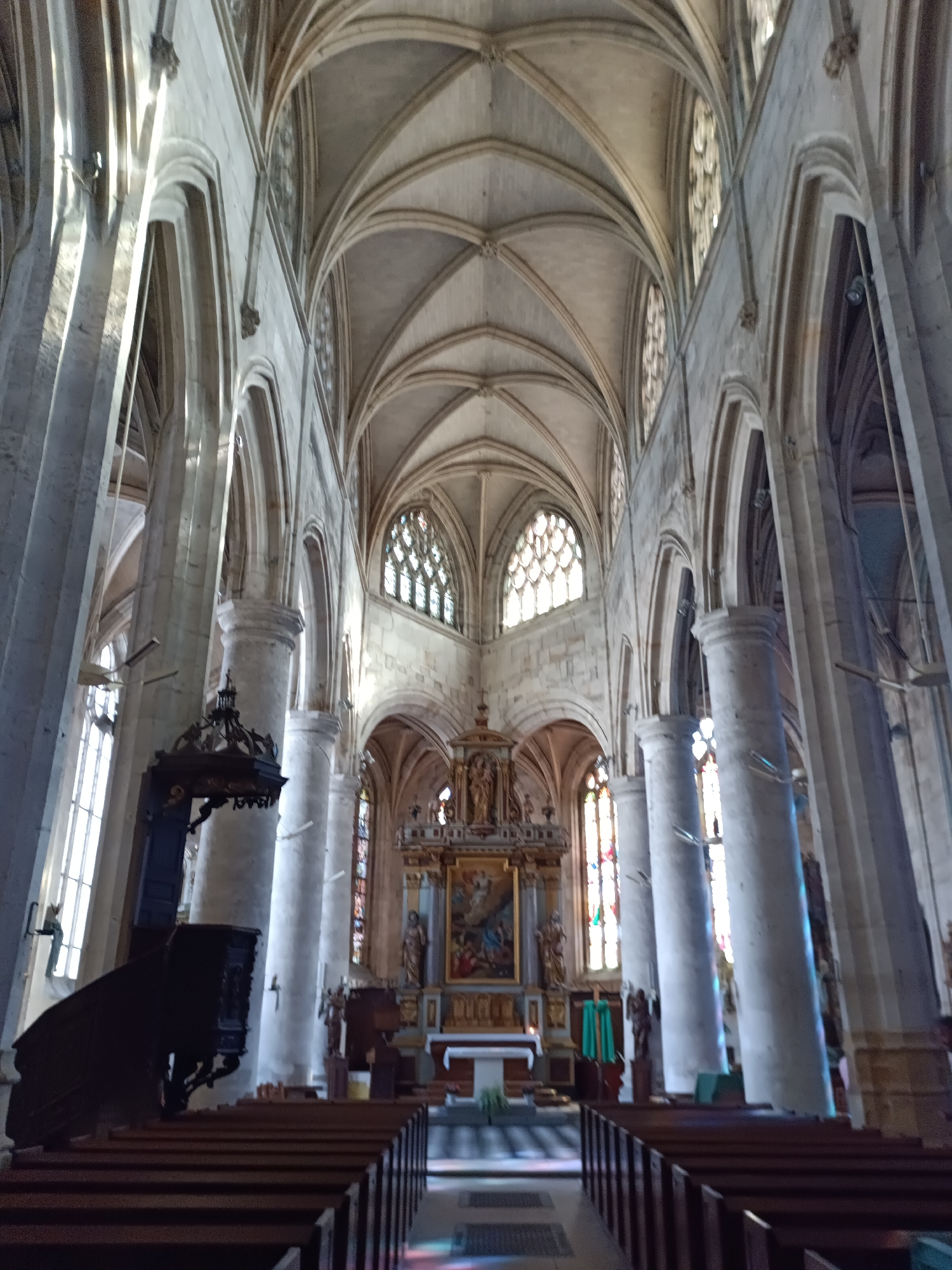 The image shows the interior of a large, old church. The architecture is Gothic, with high vaulted ceilings, pointed arches, and large stained-glass windows. The church is furnished with rows of wooden pews facing an altar at the far end. A dark wooden pulpit is visible on the left side of the image. Sunlight streams in through the windows, illuminating the stone pillars and casting colorful patterns on the floor. The overall impression is one of grandeur and solemnity. The church is Église Notre-Dame, located in Le Neubourg, France. There are no people visible in the image.