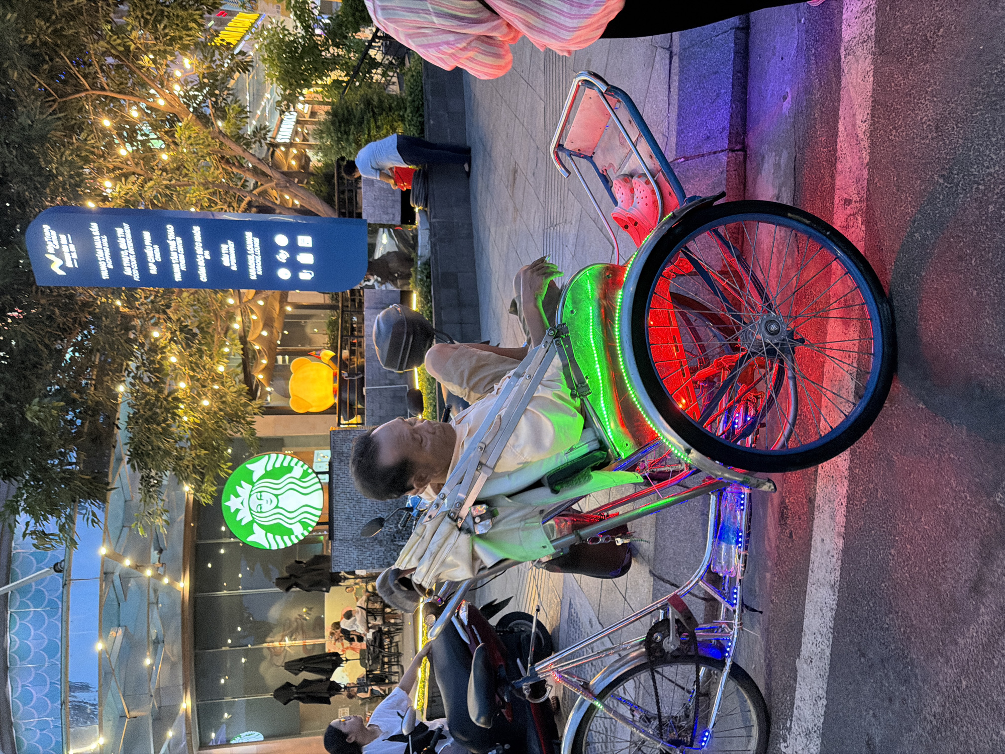 An older man of Asian descent sits in a brightly lit, three-wheeled pedicab, his eyes closed as if resting. The pedicab is adorned with vibrant green and red LED lights along its frame and wheels, casting colorful hues on the surrounding asphalt and sidewalk. A white umbrella is propped up behind him, providing shade. In the background, a large Starbucks logo glows green, and a tall blue sign lists various establishments, suggesting a commercial area in Nha Trang Ward, Vietnam. Several people are visible in the background, some seated at tables outside a building, others walking. A parked motorcycle is visible to the left of the pedicab. The scene appears to be captured during twilight, with artificial lights illuminating the area.