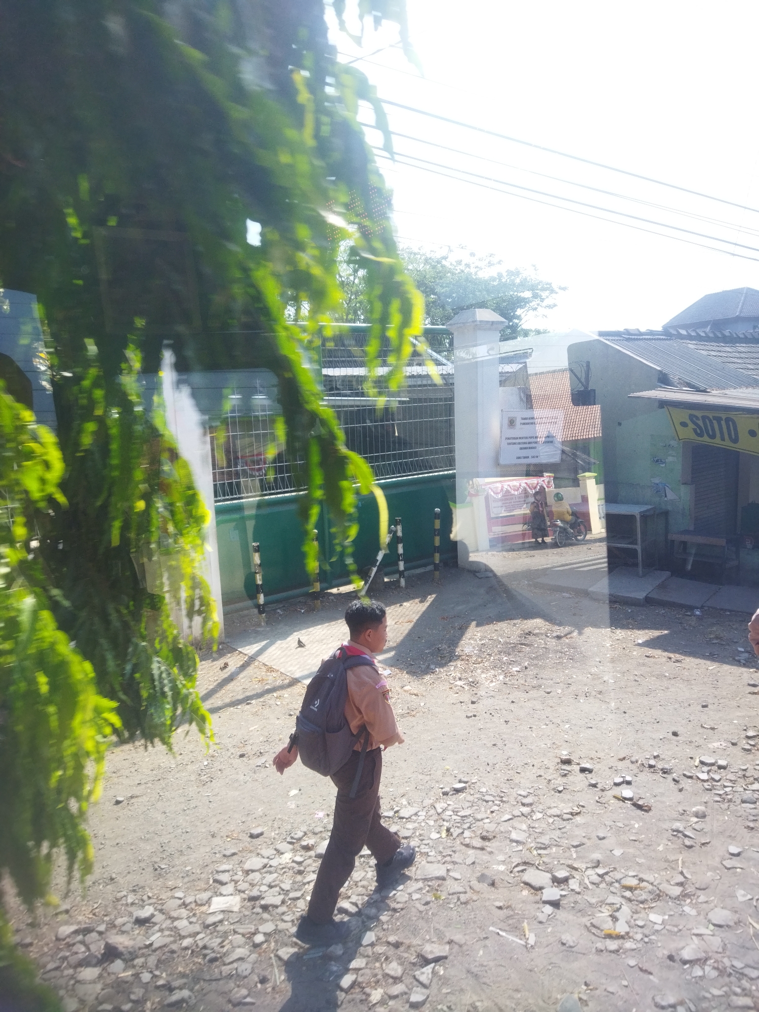 In Nambangan, Indonesia, a young boy in a brown school uniform walks on a gravel path. He wears a dark backpack over his shoulders and appears to be walking away from the viewer. The scene is illuminated by bright sunlight, casting shadows on the ground. In the background, there is a green gate with a fence, a building with a tiled roof, and a sign that reads "SOTO". Some trees and power lines are also visible. The ground is covered with small stones and dirt.