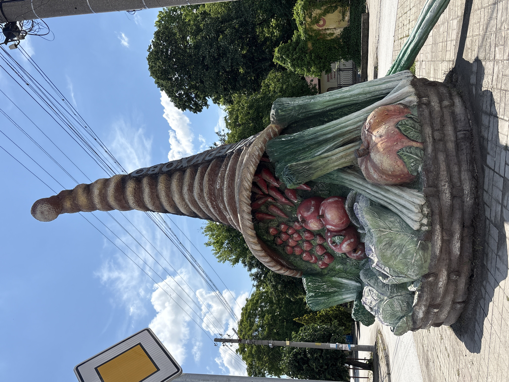 The image shows a large, sculpted cornucopia, or horn of plenty, overflowing with vegetables, including tomatoes, onions, leeks, and cabbages. The sculpture is located in the city of Biaga, Bulgaria. The cornucopia is made of a textured, weathered material, likely concrete or similar, and painted to resemble woven straw and the colors of the various vegetables. It is situated on a paved walkway near a street with overhead power lines and foliage. In the background, there are trees and possibly a building. A yellow diamond traffic sign is visible near the bottom left corner of the image. The sky is blue with scattered clouds.