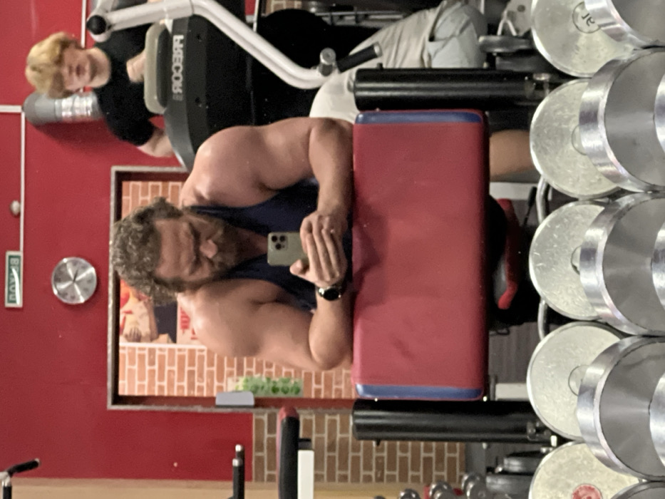 A gym setting captures a muscular man taking a selfie, positioned next to a red padded bench and a rack of silver weights. His reflection is visible in a mirror, revealing another person in the background using gym equipment. The gym's wall is red, adorned with a sign and a decorative brick pattern. The overall scene conveys a fitness-oriented environment, with the subjects engaged in physical activity.