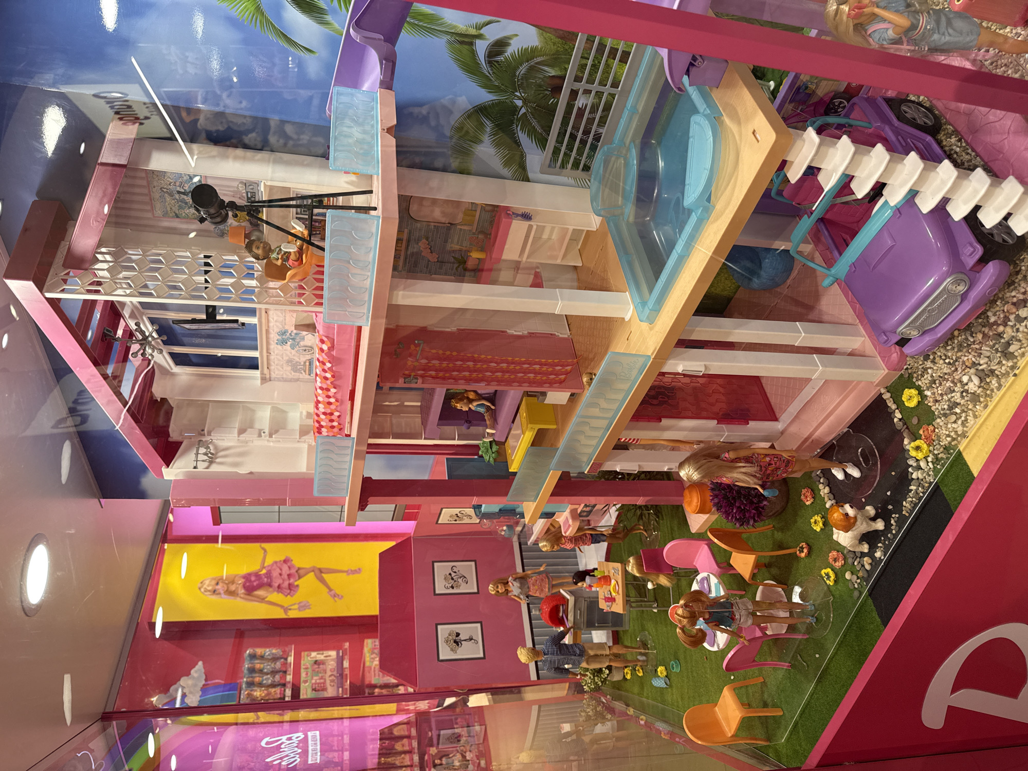 The image displays a multi-level Barbie Dreamhouse. The house features a pool, a slide, and multiple rooms with furniture and accessories. Several Barbie dolls are positioned in various areas of the house, simulating everyday activities. The background suggests an outdoor scene with palm trees. The overall setting appears to be a toy display, potentially located in a retail environment. There is no visible evidence indicating that this is located in Moscow, Russia.