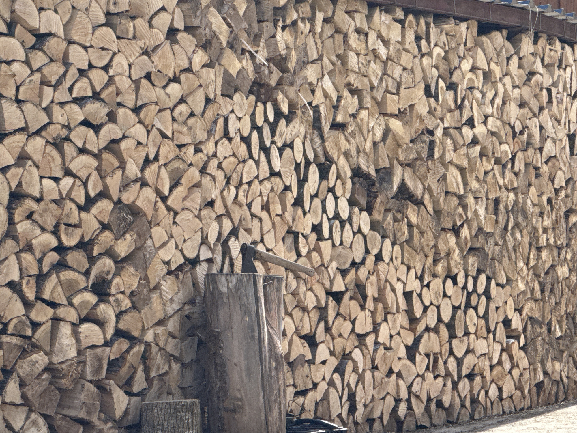 A large stack of cut firewood fills the frame, appearing to be stacked against a wall or under a covering. In the foreground, an axe is embedded in a wooden chopping block. The image does not provide enough context to determine the location in Villanova or Villanoba, Spain.
