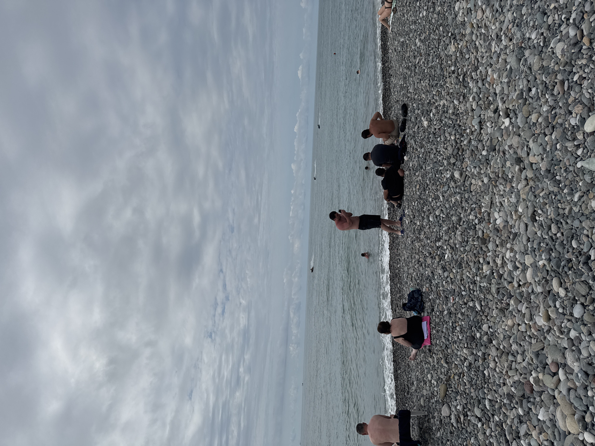 This image depicts a pebble beach in Batumi, Georgia, under a cloudy sky. The sea is calm with gentle waves lapping the shore. Several people are present, some sitting on the pebbles, while others are in the water. In the distance, jet skis are visible on the sea.
On the left side of the image, a man is sitting on a chair facing the sea. To his right, a woman in a black swimsuit is seated on a pink towel. Further to the right, a group of men are gathered on the pebbles. One man is standing and looking at his phone, while others are seated. One man in the group is wearing beige shorts. To the right of this group, another person is lying down on the pebbles.
In the water, a person is swimming closer to the shore, and further out, several jet skis are being ridden. The beach is covered in smooth, grey pebbles of various sizes. The overall atmosphere appears relaxed, with people enjoying a day at the seaside.