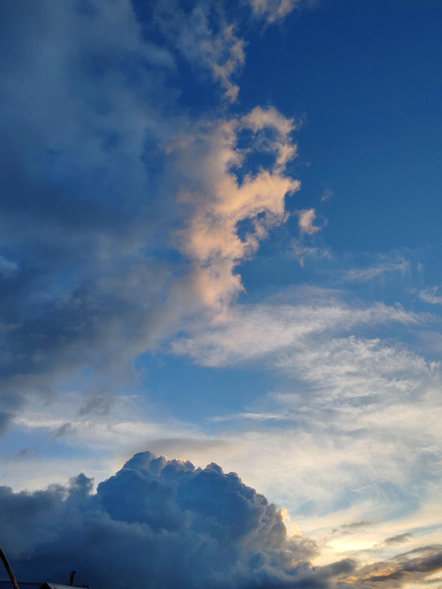 The image depicts a dramatic sky with a variety of cloud formations above a portion of what appears to be a building or structure in the lower left corner. The sky is a deep blue, transitioning to lighter shades at the horizon where the sun is setting or rising, casting a warm, golden glow on some of the clouds. Several large, dark cumulonimbus clouds dominate the lower half of the image, with lighter, wispy cirrus clouds streaking across the upper portion. One prominent cloud formation in the center is illuminated by the sun, showing shades of pink and orange. The scene suggests a change in weather. The provided location of Чухлэм, Russia, indicates an outdoor, potentially rural or suburban setting. No people or specific actions are visible in the image.