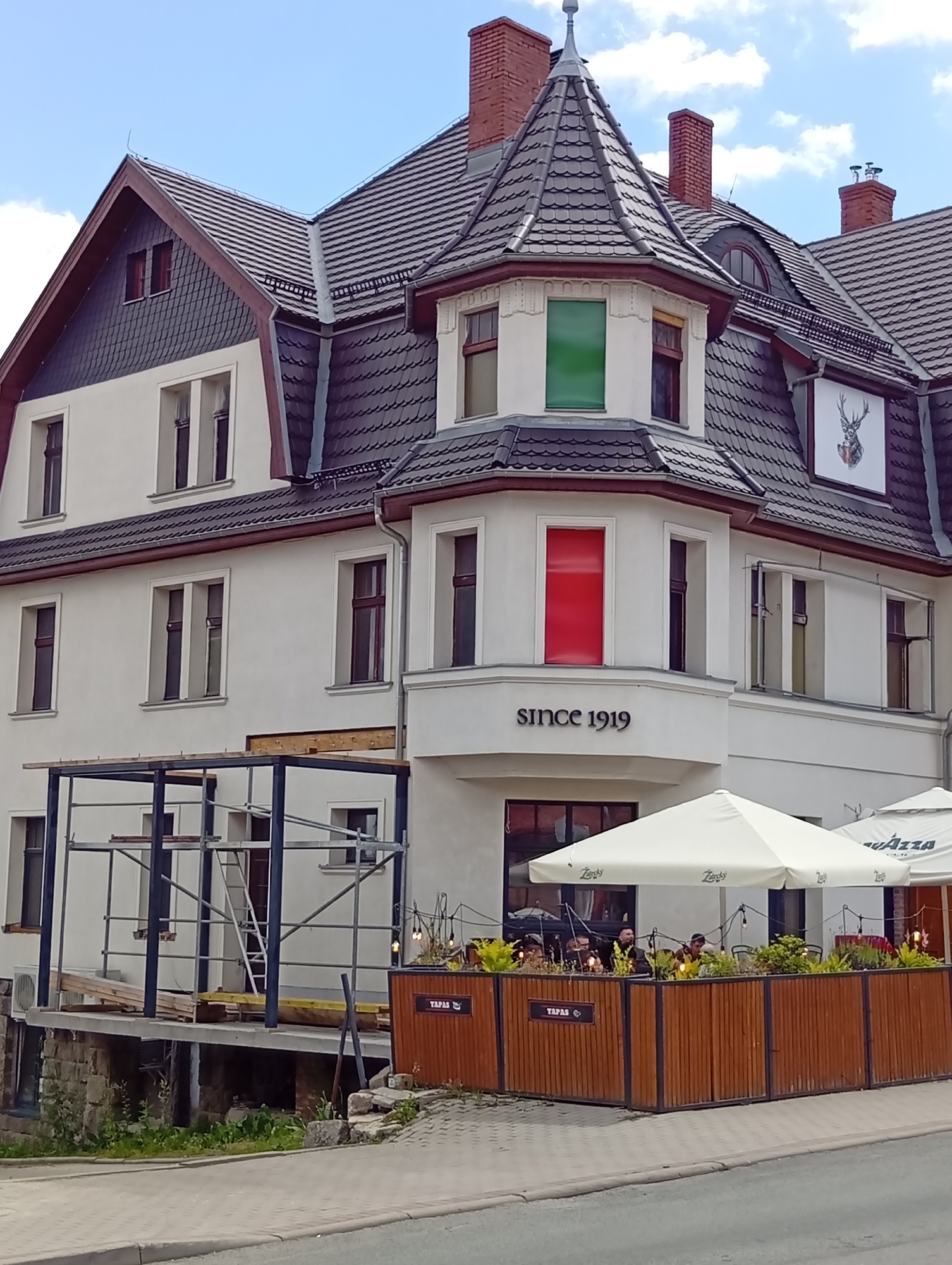 The image shows a white building in Karpacz, Poland. The building has a dark tiled roof and several windows. On the front of the building, there is a sign that says "SINCE 1919". There is also a white umbrella and some outdoor seating in front of the building. A deer's head is depicted on a sign on the side of the building. The sky is blue with some clouds. The scene appears to be a daytime shot. There are no people clearly visible in the image, though there are silhouettes of people sitting at tables under the umbrellas. The building appears to be a restaurant or cafe. There is scaffolding on the side of the building indicating construction or renovation.