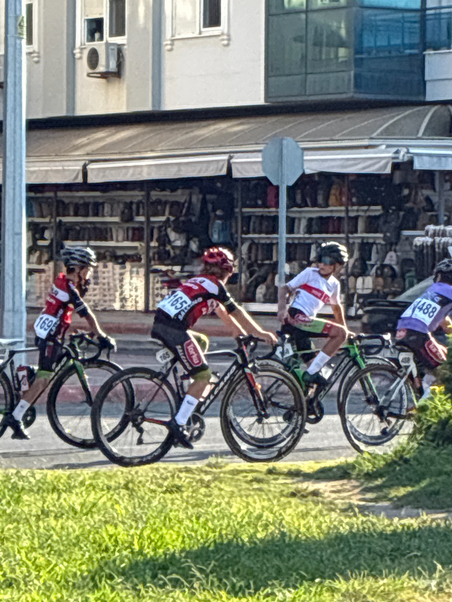 Four cyclists in racing gear, with helmets and cycling shoes, are participating in a race on a paved road. The cyclists are numbered: 1695, 165, and 488. The cyclist with number 165 is wearing a red, white, and black jersey and green shorts. The cyclist with number 488 is wearing a purple jersey and black shorts. The background shows a building with shops and an awning, a street sign, and a grassy area. The scene appears to be in Alanya, Turkey, judging by the architecture and environment. The image captures a moment during the race, with the cyclists in motion, their bodies leaning into the turns of the road. The bright sunlight casts shadows on the ground, indicating it is daytime.