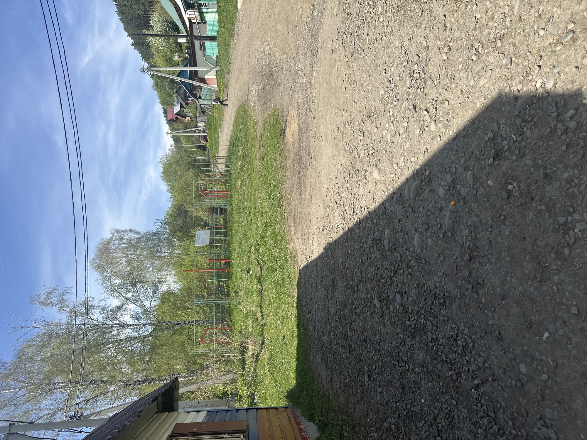 The image shows a partly sunny day in Gorno-Altaysk, Russia. A gravel road runs diagonally across the frame, with a dark shadow cast onto its right side. To the left of the road, there is a grassy area with trees and a metal playground structure. In the background, residential buildings and trees are visible. The sky is blue with scattered clouds. Power lines are present in the upper left quadrant. A portion of a building with wood siding is visible in the lower-left corner. A single person is walking in the distance on the road.