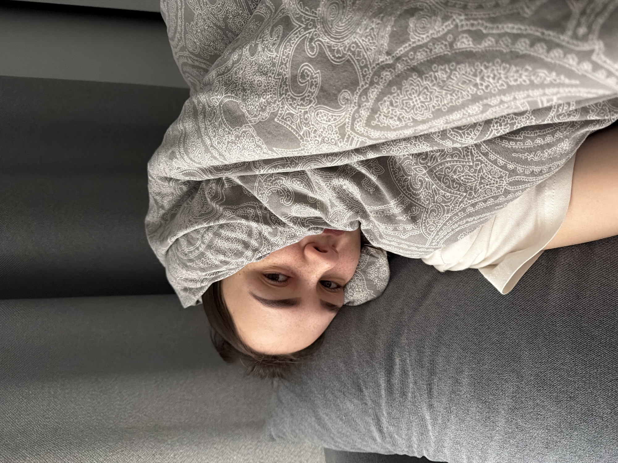 A person lies on a gray surface, possibly a sofa or bed, covered with a gray patterned blanket. Only their face is visible, peeking out from under the blanket. The person has short brown hair, fair skin, and is wearing a white shirt. The image appears to be taken indoors, possibly in a residential setting. There is no specific evidence to confirm the location as Chaikovsky, Russia.