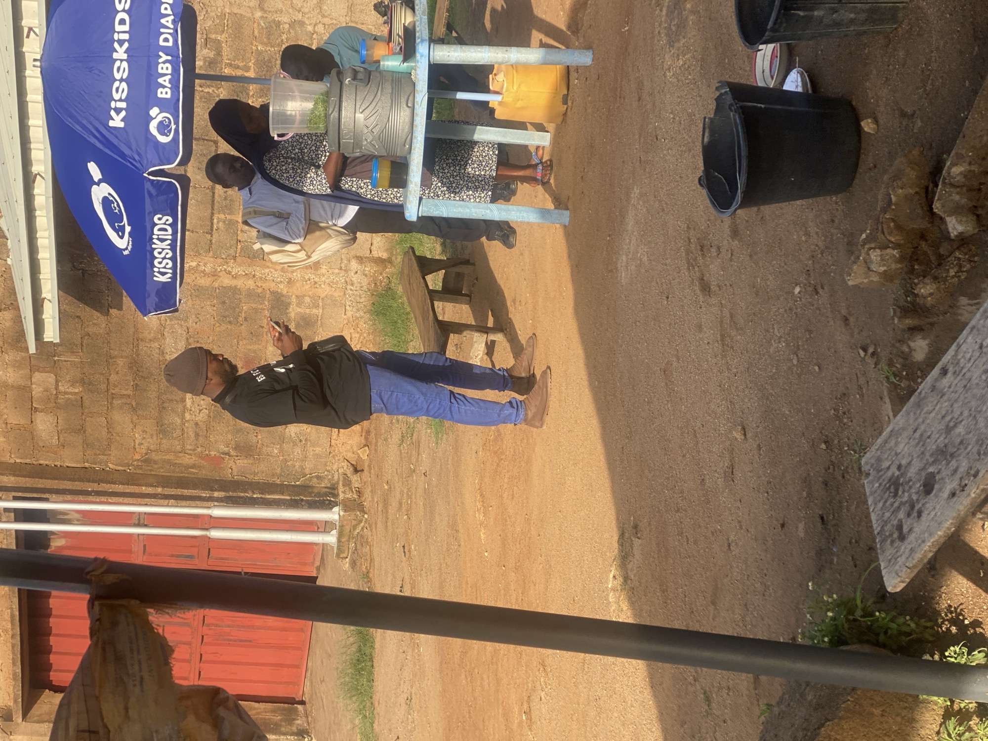 The image captures a street scene in Jos, Nigeria, under bright daylight. A man wearing a beanie and a black t-shirt stands looking at his phone, dressed in blue jeans and brown shoes. To his right, a woman in a patterned dress and hijab stands near a table laden with containers and cups. Behind her, a man in a light-colored shirt and another man in a light blue shirt are also present. A blue umbrella with "Kisskids BABY DIAPER" branding is set up, providing shade. The ground is a dusty, sandy earth, with a wooden bench visible to the left of the table. In the foreground, part of a dark pole and a wooden plank are visible. The background features a building with a red door and a textured stone wall. Several black buckets and a yellow container are scattered on the ground.