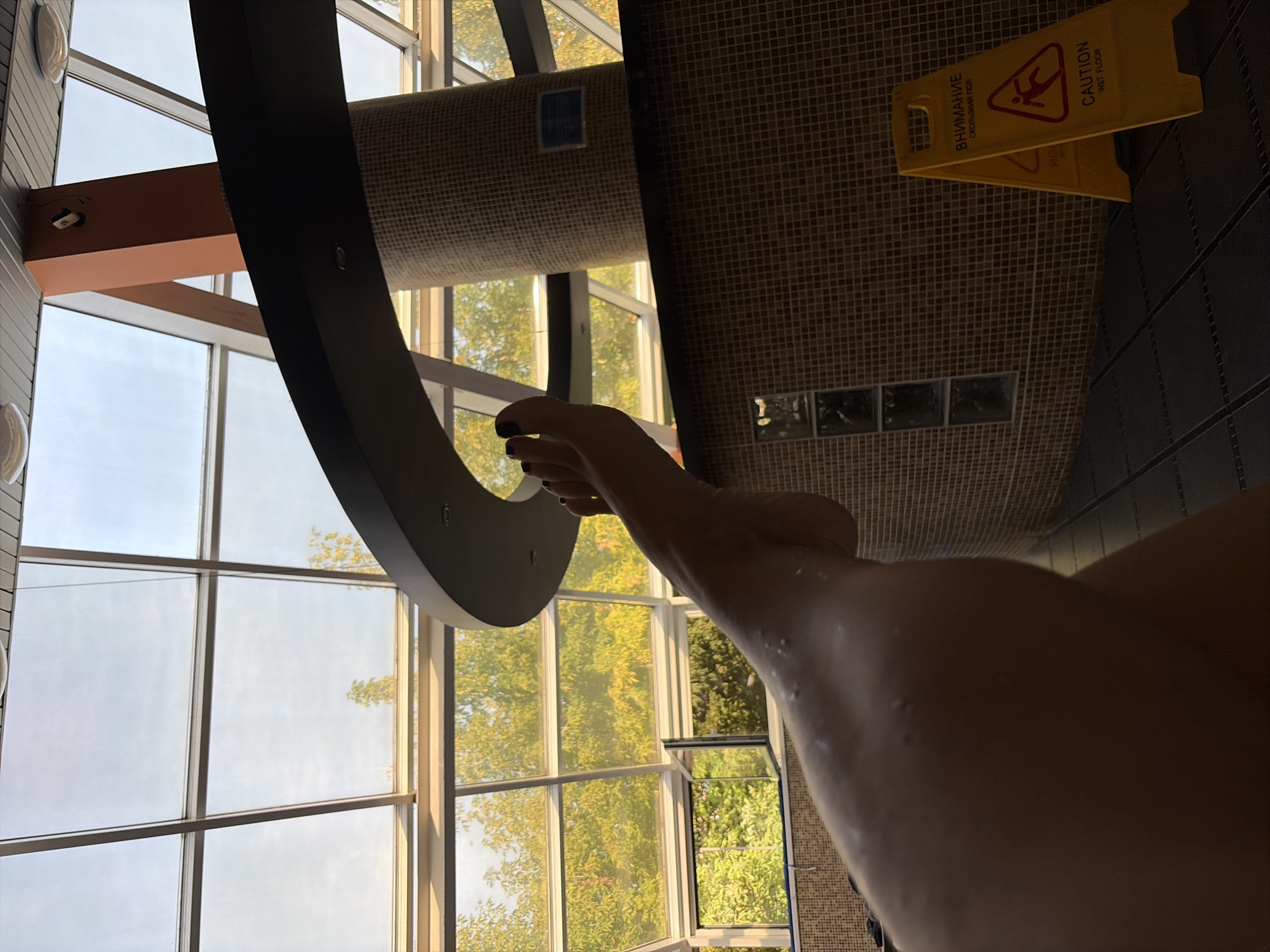 A person's leg and foot, with dark nail polish and water droplets, are positioned in the foreground, angled towards the left of the frame. The individual appears to be near a pool or spa area, as indicated by the wet leg and a yellow "Caution: Wet Floor" sign visible in the lower right corner. The background reveals a large, glass-paneled ceiling, allowing natural light to illuminate the interior. Through the glass, trees with yellow and green foliage are visible, suggesting an outdoor setting or an area with a view of nature. The architecture includes a large, tiled cylindrical pillar and a curved dark structure overhead. The overall scene depicts an indoor recreational space with a view of the outdoors, possibly a public pool or a spa within Moscow, Russia.