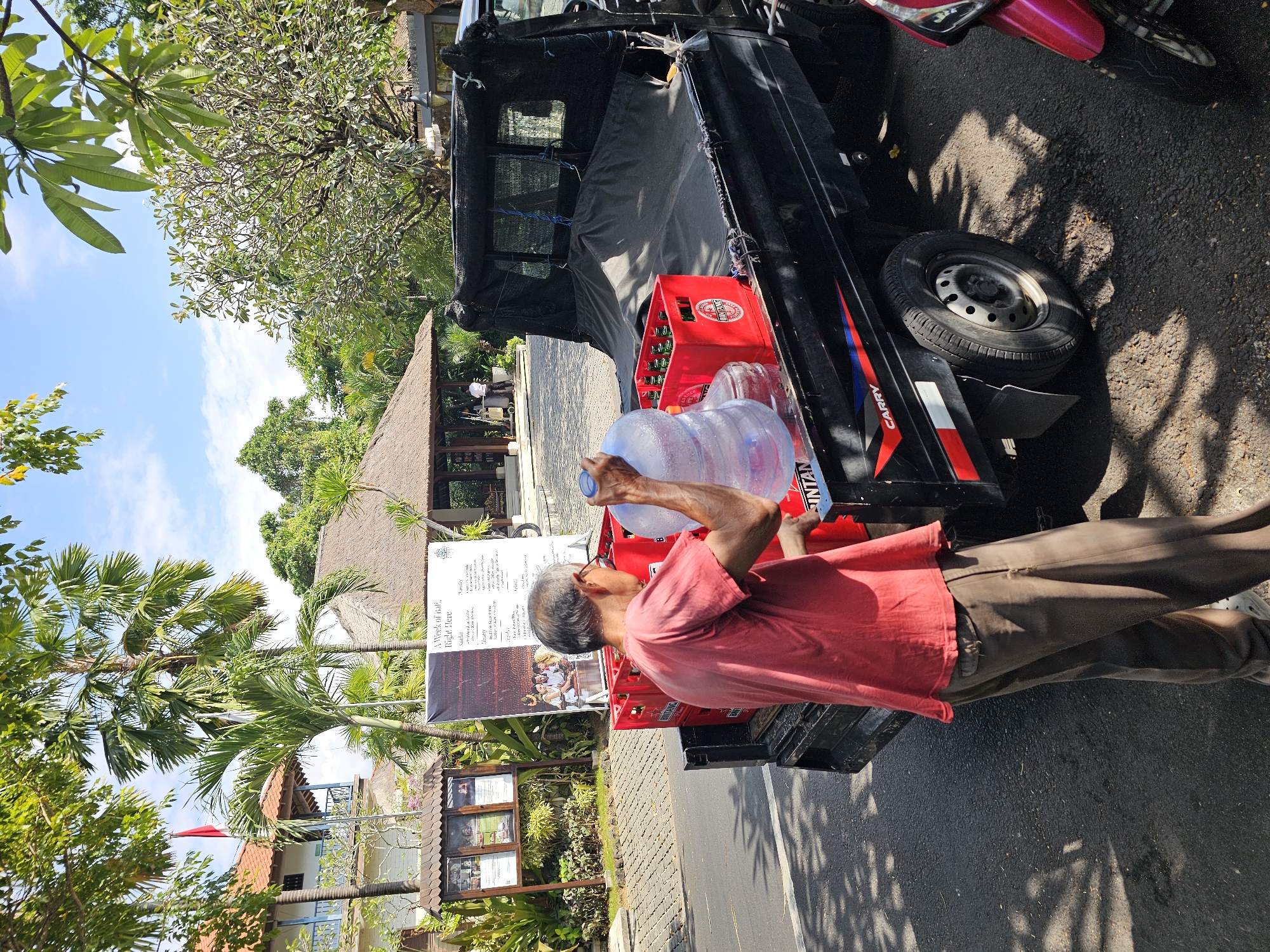 A man, wearing a red t-shirt and brown pants, is unloading large water jugs and red crates of bottled beverages from the back of a black utility truck. The truck is parked on a paved road in front of a resort-like building with thatched roofs and palm trees. There is a sign with images of people and text visible behind the man. The scene appears to be in Denpasar, Indonesia, given the tropical foliage and architectural style. Sunlight casts shadows on the ground.