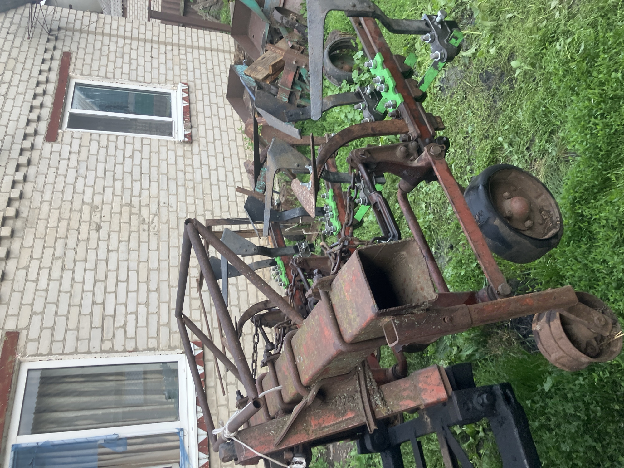 In the village of Vovkivka, Ukraine, a rusty, old agricultural machine, possibly a plow or cultivator, sits on a grassy field in front of a light brick building with white-framed windows. The machine is complex, with many metal parts, chains, and wheels. Some parts of the machine are painted red, and some components are bright green. The building appears to be a house or outbuilding, with two visible windows. The overall scene suggests a rural, possibly agricultural setting.