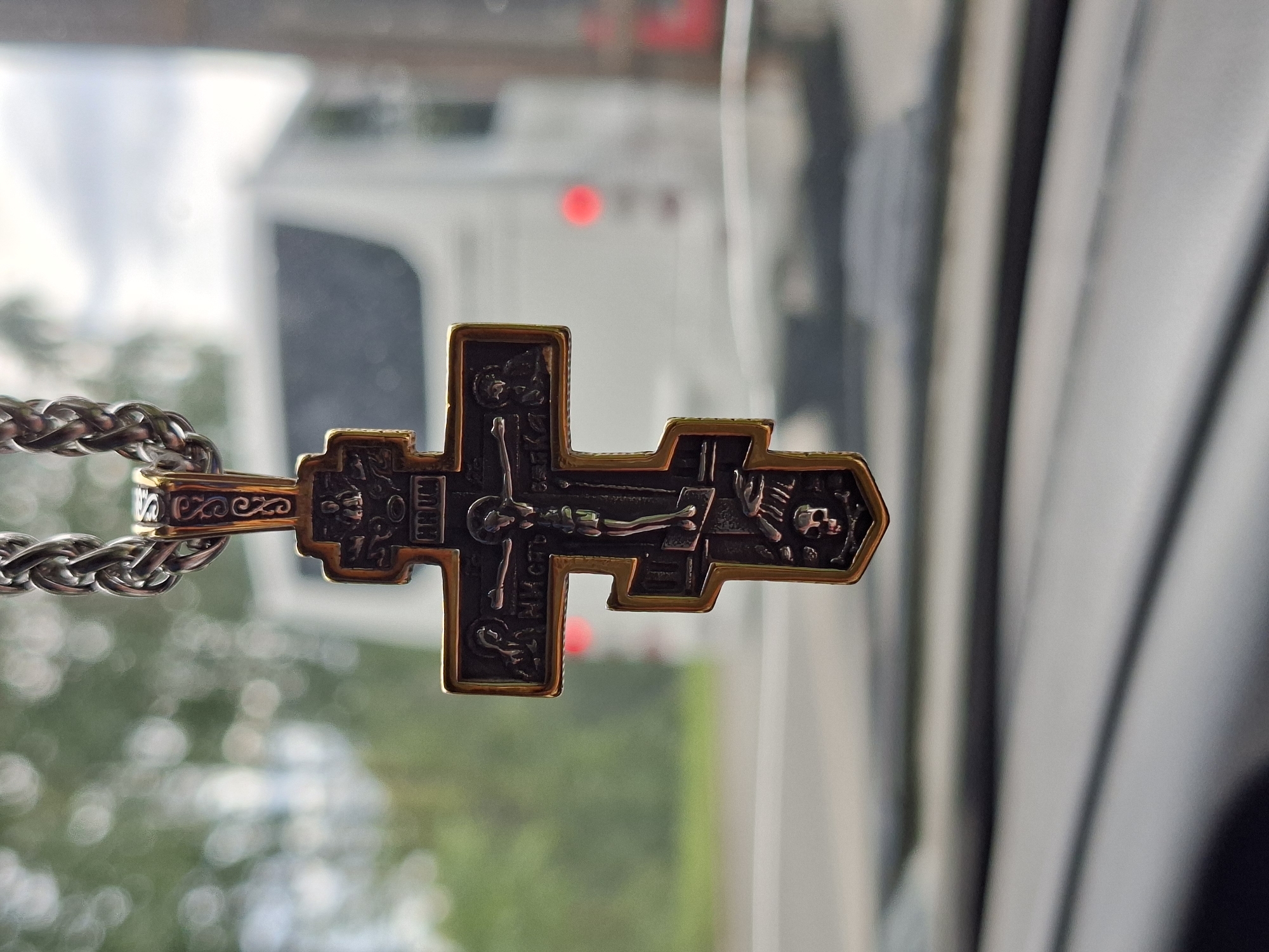 A close-up, vertical shot focuses on a decorative Orthodox Christian cross hanging from a silver chain. The cross, with a gold trim, features an image of Jesus Christ crucified at the center, along with other religious figures and symbols, including a skull at the bottom. The cross is positioned in the foreground, obscuring the view of the interior of a vehicle, likely a car, where the photograph is taken from. The background is blurred and depicts an outdoor scene with a white bus or similar vehicle on a road, suggesting a daytime setting. The environment appears to be a city, potentially Chaikovsky, Russia, based on the context provided.