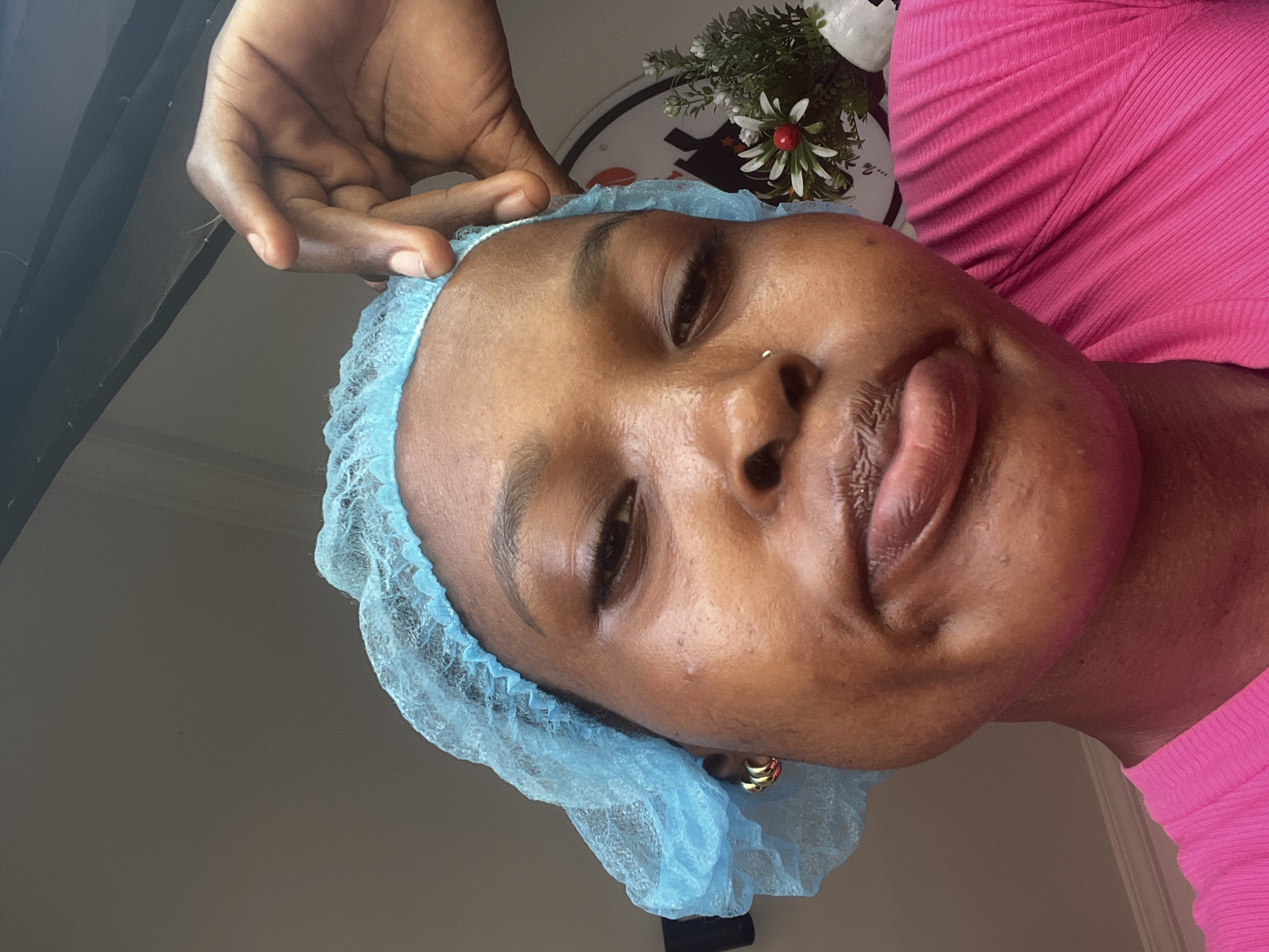 A close-up shot features a person wearing a light blue surgical cap and a pink shirt. A hand is visible near the top of the image, seemingly adjusting the cap. The person has dark hair and skin. The background includes a wall, part of a decorative item, and a window. The image's location is indicated to be in Lekki, Nigeria, although no details in the image itself can verify this.