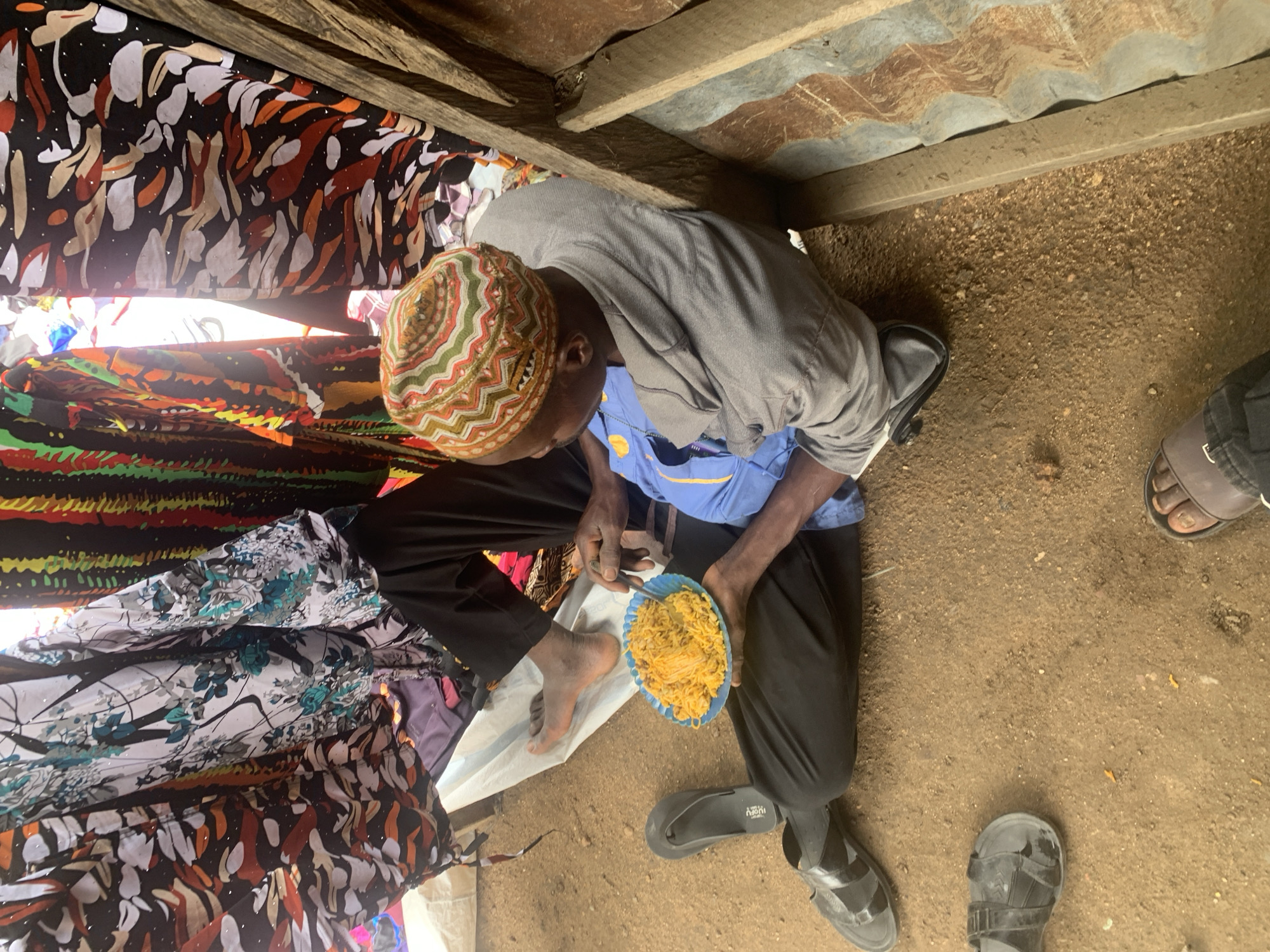 The image depicts a person sitting on the ground in what appears to be an outdoor setting in Project, Nigeria. The individual is wearing a grey hooded sweatshirt, a blue shirt underneath, black pants, and a multi-colored kufi cap. They are holding a blue plate filled with yellow food, identified as rice, and are in the process of eating it with a fork. The person is barefoot, wearing black sandals on their left foot, and other sandals are visible on the ground nearby, along with a second person's foot wearing brown sandals. The background features colorful fabrics hanging, suggesting a market or a domestic setting. The ground is composed of dirt and small stones. Wooden planks and corrugated metal are visible on the right side of the frame, indicating a rustic structure. The lighting suggests it is daytime.