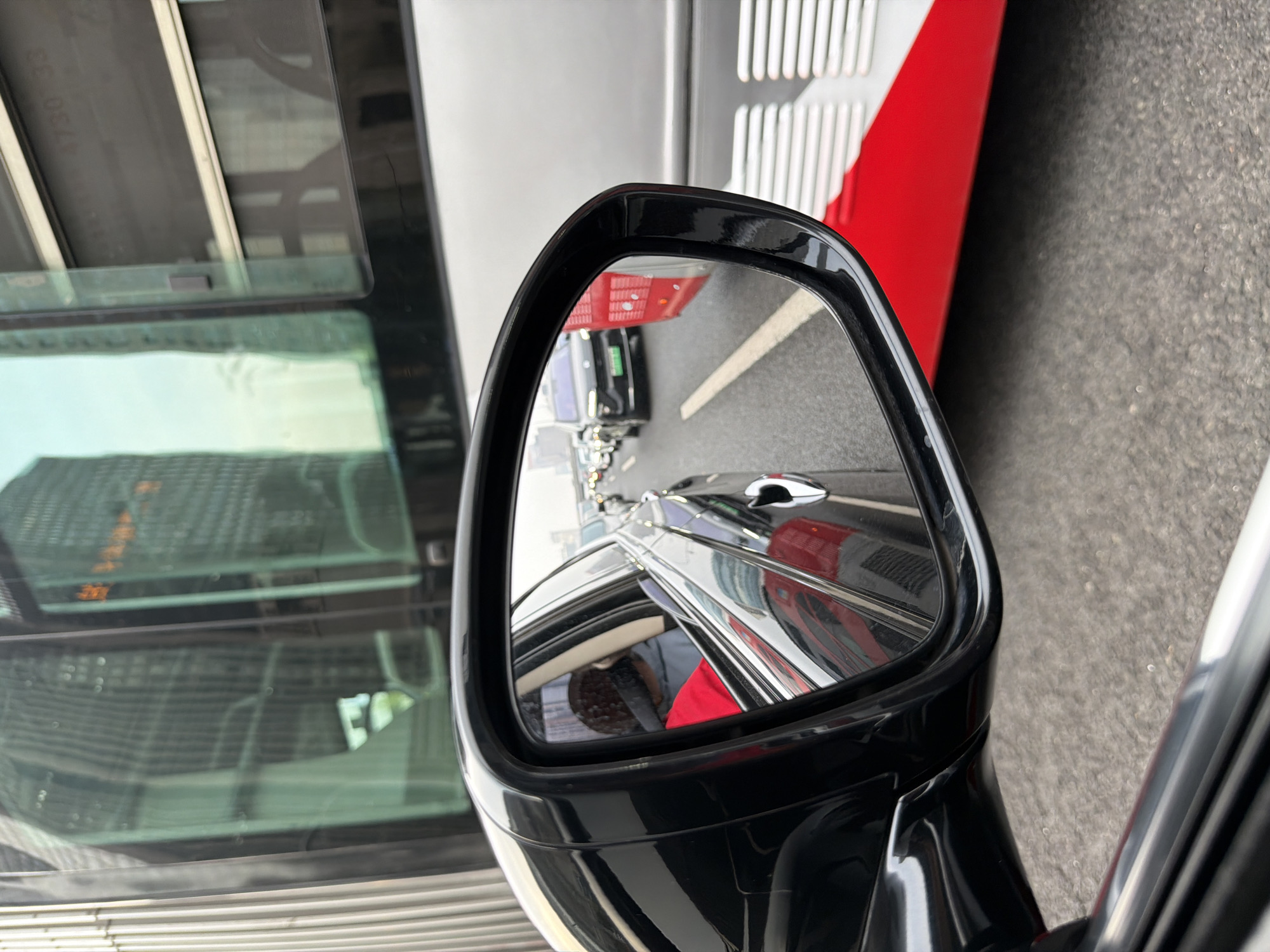The image shows a driver-side rearview mirror of a car. The mirror's reflection reveals a row of vehicles lined up on a road, suggesting traffic congestion. Buildings are visible in the background, suggesting an urban setting. A part of a red and white vehicle is visible on the right side of the rearview mirror. Another building with a glass facade is to the left. Based on the context, the location is possibly in Chaoyang District, China.