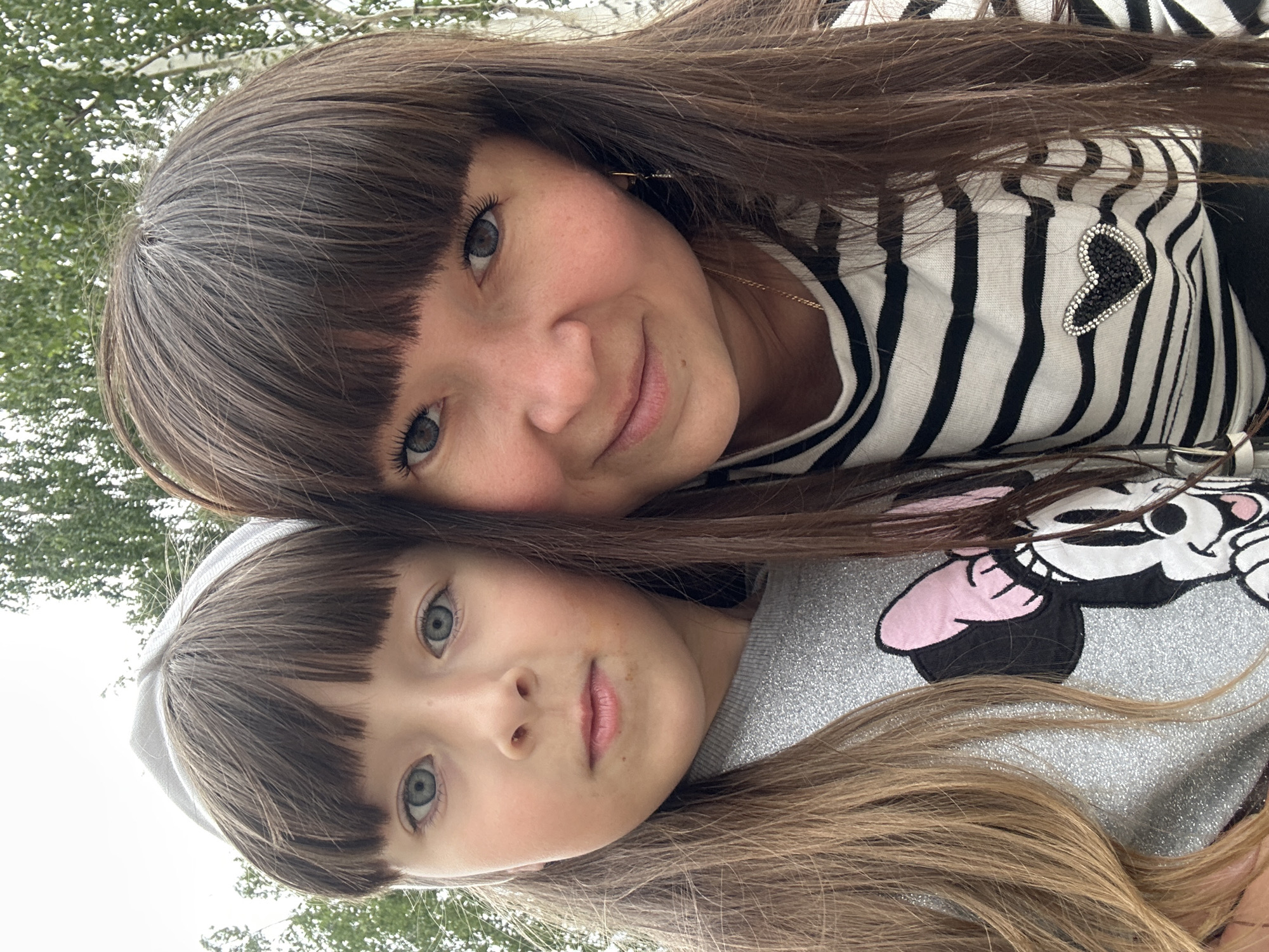 The image is a selfie taken outdoors, with trees and a bright sky in the background. In the foreground, two females are visible. The female on the left, likely a child, has light brown hair with bangs and is wearing a grey, sparkly sweater with a Minnie Mouse graphic. She has blue eyes and is looking directly at the camera. The female on the right, likely an adult, has dark brown hair with bangs and is wearing a black and white striped shirt with a heart-shaped embellishment. She is smiling and her eyes are also blue. They appear to be close together, suggesting a familial relationship. The overall setting suggests a casual outdoor activity. There is no specific indicator of the location in the city of Chaikovsky, Russia.