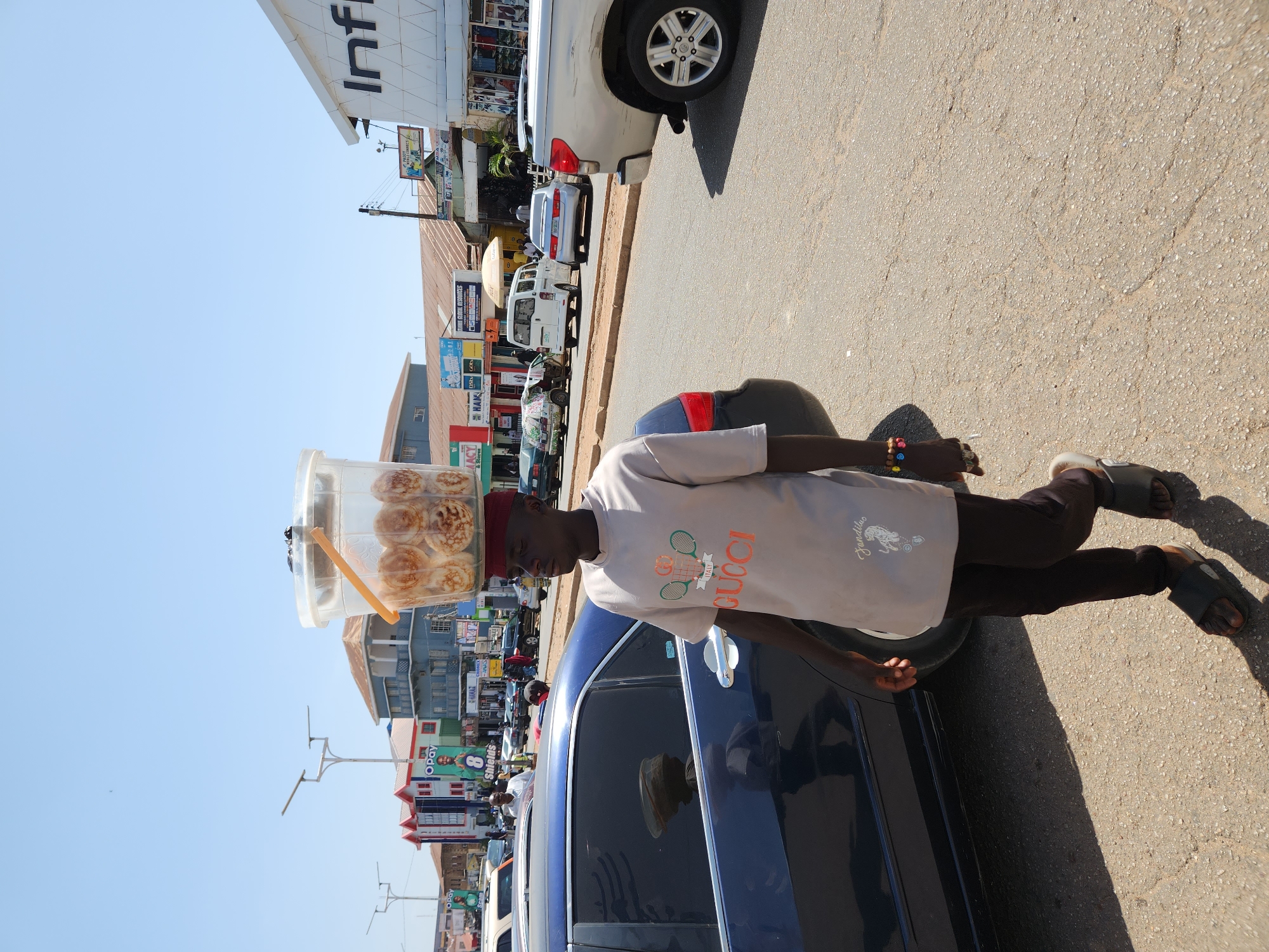 A man balances a container of baked goods on his head in a city. He is wearing a beige shirt, brown pants, sandals, and a red fez. The container on his head appears to be clear plastic and filled with small, round pastries. He stands next to a dark-colored car on a paved street. The background features various buildings, including one with the sign "Infi" visible. Multiple vehicles are parked along the street. The sky is clear and blue.