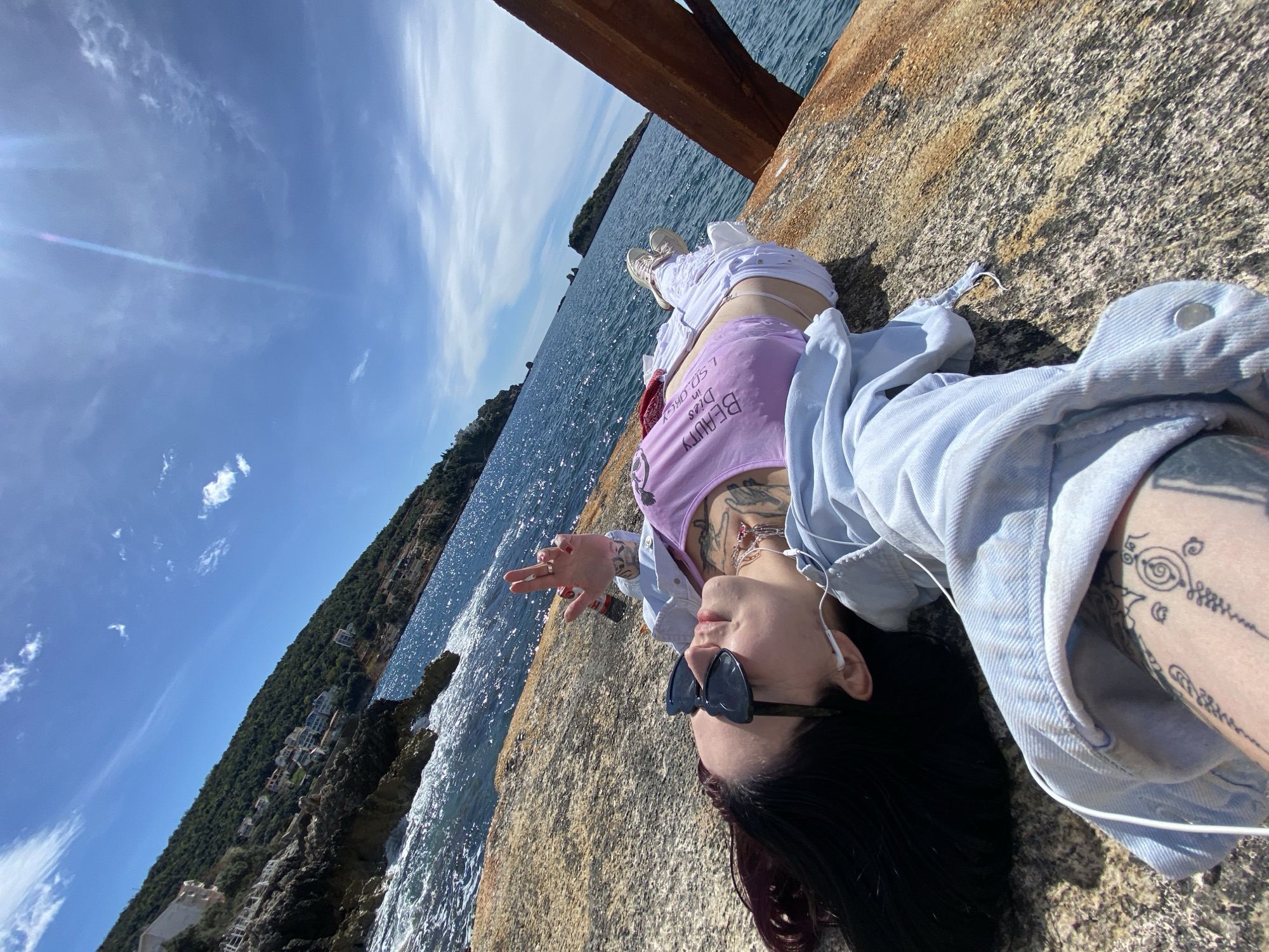 A woman lies on a concrete structure adjacent to the sea. The structure, which appears to be a pier or retaining wall, shows signs of weathering. The woman has dark hair, sunglasses, and a lavender tank top. She is wearing a light blue denim jacket. Tattoos are visible on her arms and chest. One hand is raised in a "peace" sign. She is wearing earbuds with white cords. The sea is blue, with visible waves. A wooded coastline stretches along the horizon. A rusty metal beam overhangs the scene. The location is identified as the city of Bar, Montenegro.