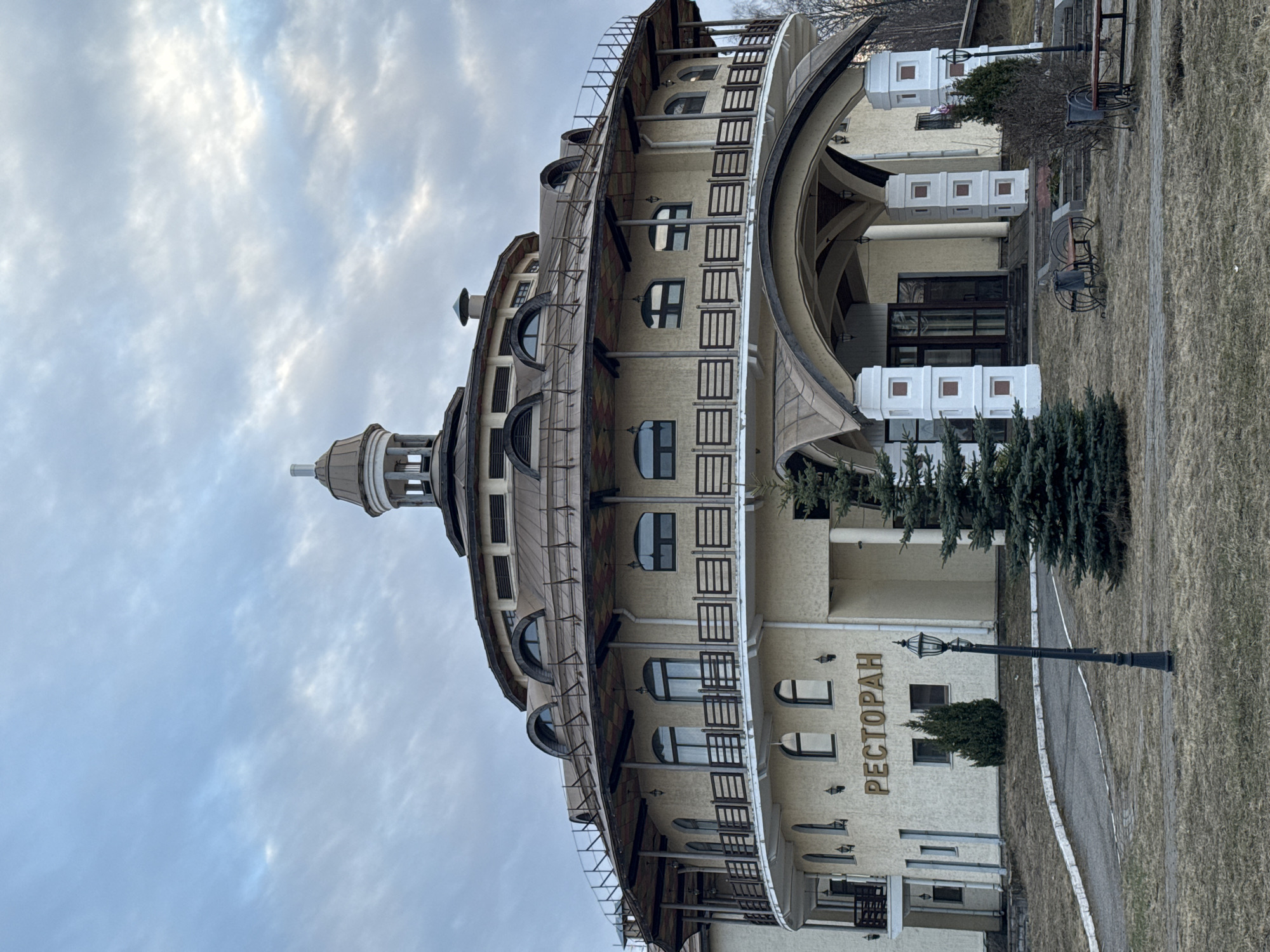The image shows a view of a cylindrical building in Ново-Никольское, Russia. The building is light-colored, featuring multiple floors with arched windows and decorative railings. A rounded roof topped with a turret-like structure sits atop the building. A sign reading "РЕСТОРАН" is visible on the lower portion of the building. Landscaping including evergreen trees and a lamp post is present. The sky is overcast with clouds. A paved walkway is nearby.