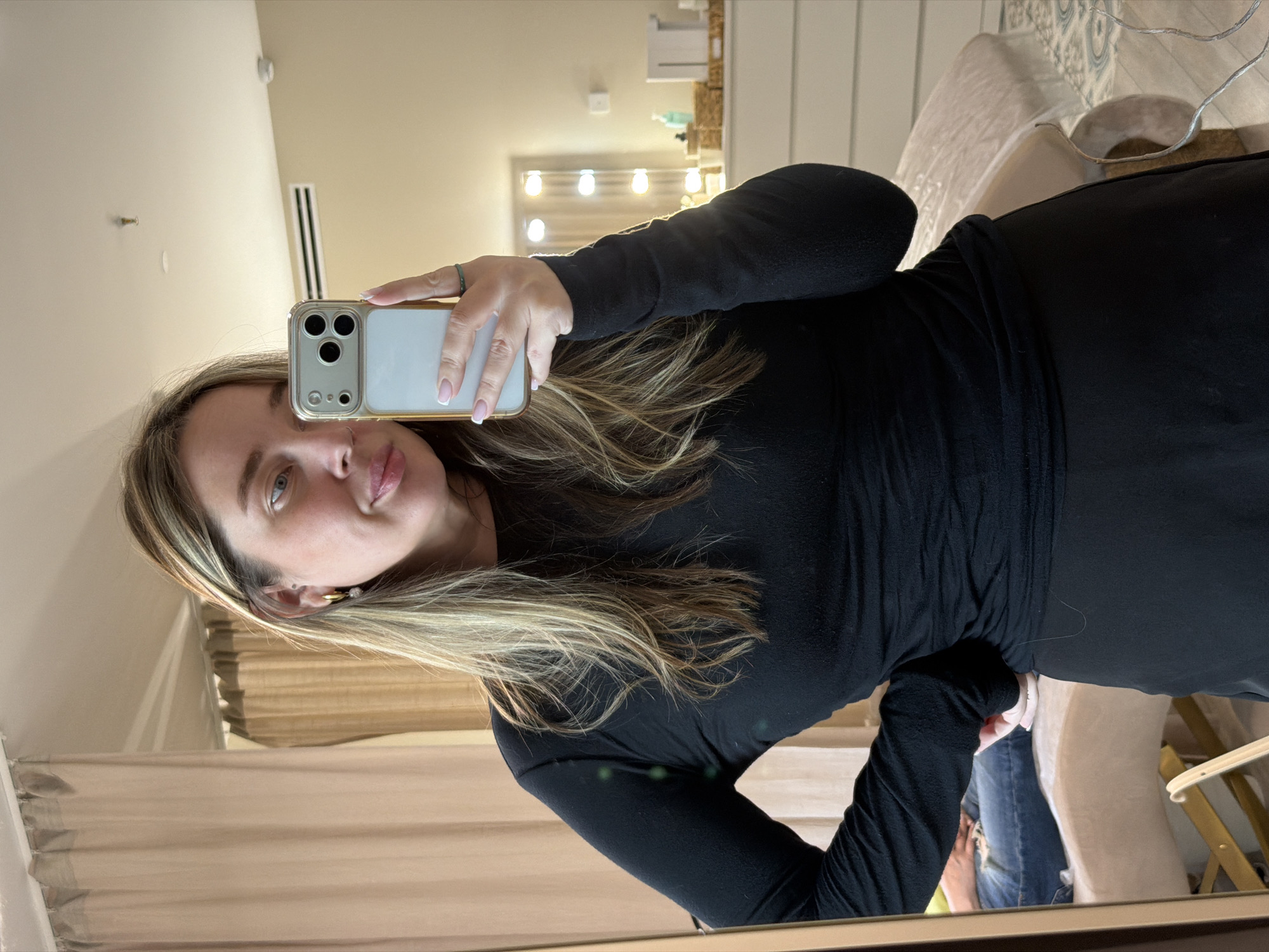 A woman is taking a selfie in a mirror. She is holding a white smartphone with a gold-colored case. She is wearing a black long-sleeved dress. Her hair is blonde with brown roots. The background includes curtains, a vanity with lights, a chair, and a patterned surface, likely a rug or cushion. The location in the city of Dolgoprudny, Russia, cannot be confirmed from the image.