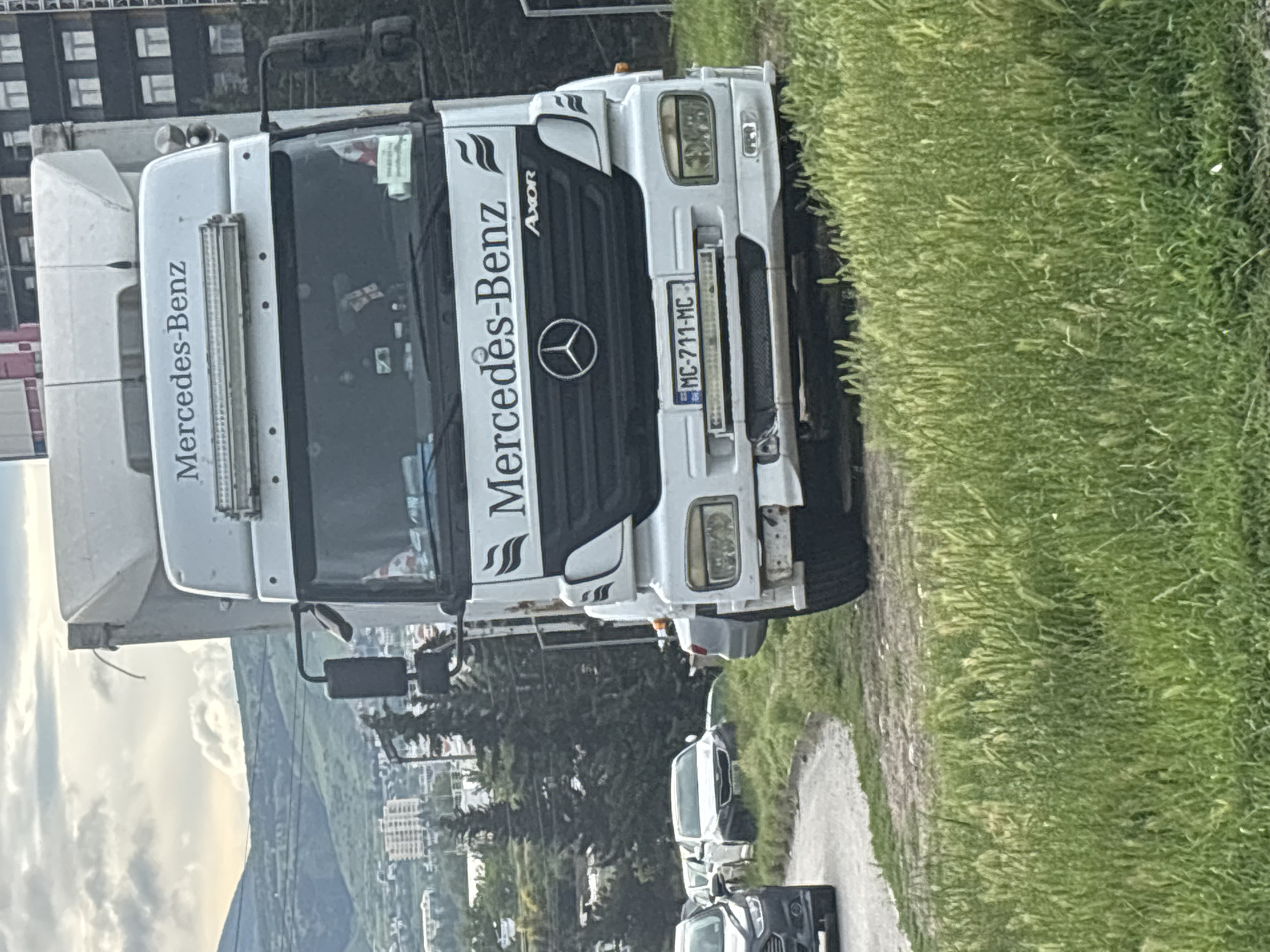 A Mercedes-Benz Axor truck with license plate "MC-711-MC" is parked on a road next to a grassy area. The city of Tbilisi, Georgia, is visible in the background, including buildings, a mountain, and trees. Several cars are parked behind the truck. The photo appears to have been taken from a slightly elevated position.