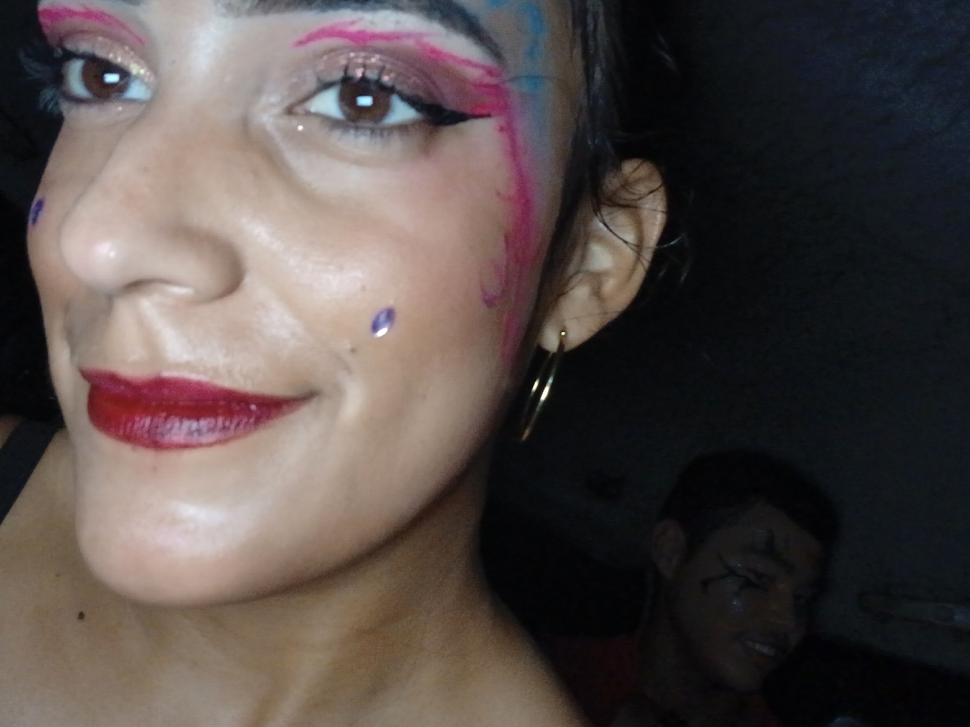 A close-up photograph shows a woman with colorful makeup on her face and around her eyes. She has pink and blue streaks near her hairline and around her eyes, a purple gem-like sticker on her cheek, and she wears a gold hoop earring. She is also wearing red lipstick. The image appears to have been taken indoors, possibly in a car, as evidenced by the blurry presence of a person in the background with dark hair and painted markings on his face. Specifics about the location in Playa El Agua, Venezuela, cannot be determined from the image. The lighting conditions appear low, which may account for the shadows and blurry background.
