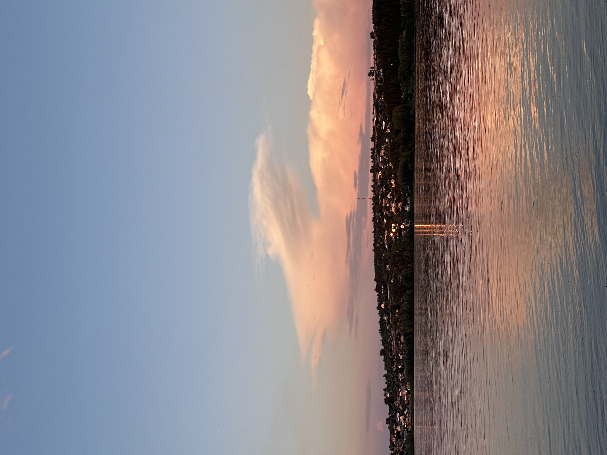 The image depicts a serene sunset over a body of water, likely the Kama River, in Chaikovsky, Russia. The sky is a gradient of soft blues and pinks, illuminated by the setting sun, with scattered clouds catching the warm light. Below the sky, a low-lying town is visible along the riverbank, characterized by numerous residential buildings nestled amongst trees. The water in the foreground is calm, with gentle ripples reflecting the colors of the sunset and the lights of the town. The overall atmosphere is peaceful and tranquil.