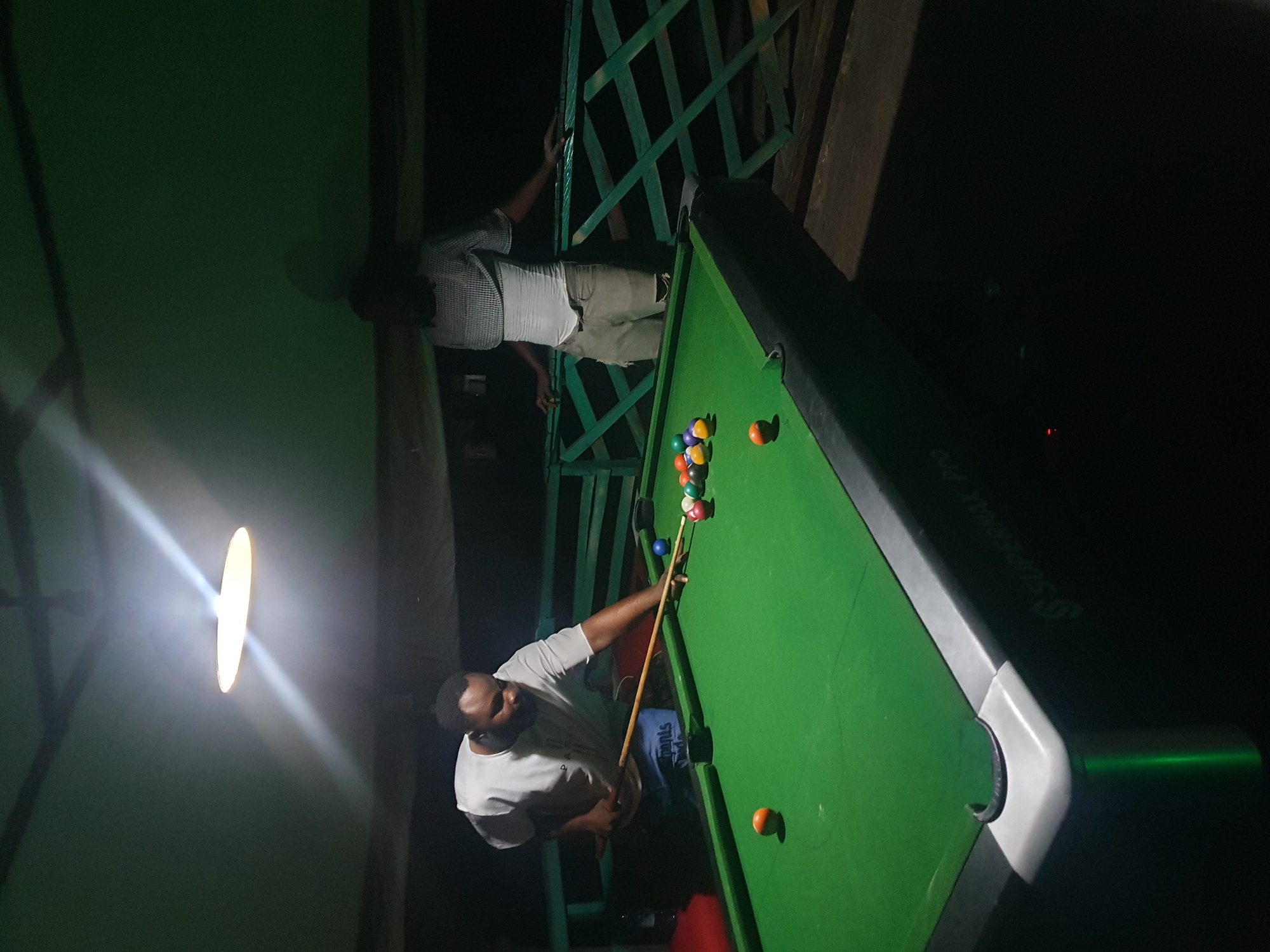 In a dimly lit room, possibly a bar or recreation center, in Lokoja, Nigeria, two men are engaged in a game of billiards. A man on the left, wearing a white t-shirt and blue shorts with writing, is in the process of striking a billiard ball with a cue stick. He is positioned over the green billiard table, which is illuminated by a overhead light. The billiard table is set up with several colored balls. Another man, dressed in a plaid shirt and light-colored pants, stands to the right, leaning on a green railing. The environment appears to be an outdoor or semi-outdoor space, with a glimpse of a wooden ceiling and green railings visible. The overall atmosphere is casual and social, with the focus on the game of billiards.