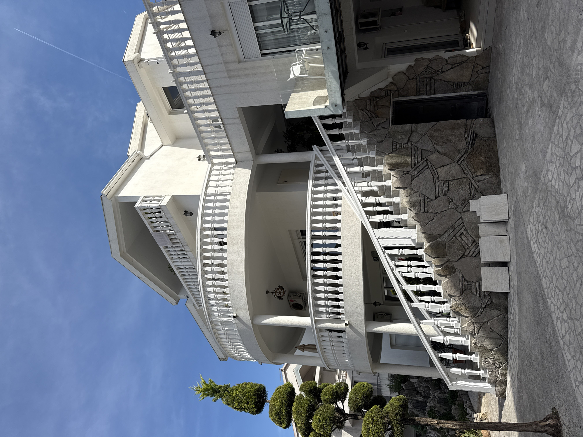 The image depicts a multi-story, white building located in Budva, Montenegro, as suggested by architectural styles common in the region. The building features curved balconies with white balustrades. A stone facade element is present near the entrance. Potted plants and landscaped shrubbery adorn the front. The sky is clear and blue. Construction materials are stacked against the side of the building.