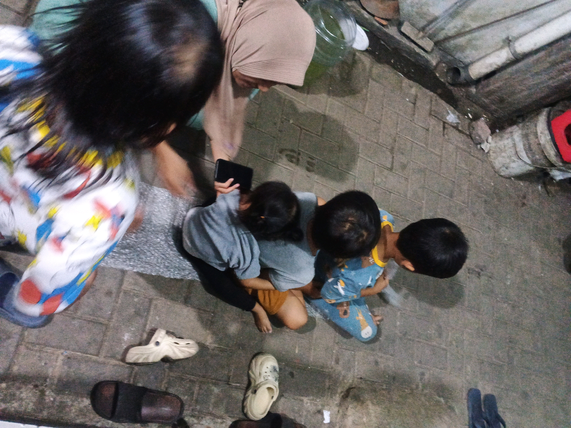 A group of four people, including at least three children, are gathered on a paved outdoor surface in Kadu, Indonesia. The perspective is from directly overhead. In the foreground, a child wearing a colorful shirt with red, blue, yellow, and white patterns is partially visible on the left. To their right, a woman wearing a light brown hijab is looking down at a smartphone held by another person, whose hand is visible. In the center and right of the frame, three children are seated or kneeling together. One child is wearing a blue patterned outfit with cartoon characters and a yellow collar. Another child is wearing a grey shirt and orange shorts, and a third child is wearing a grey shirt. Scattered on the ground are several pairs of footwear, including white clogs, dark-colored slides, and blue flip-flops. The environment appears to be an urban outdoor area with brick-like paving stones and some pipes visible in the background, suggesting it might be a residential or communal space.
