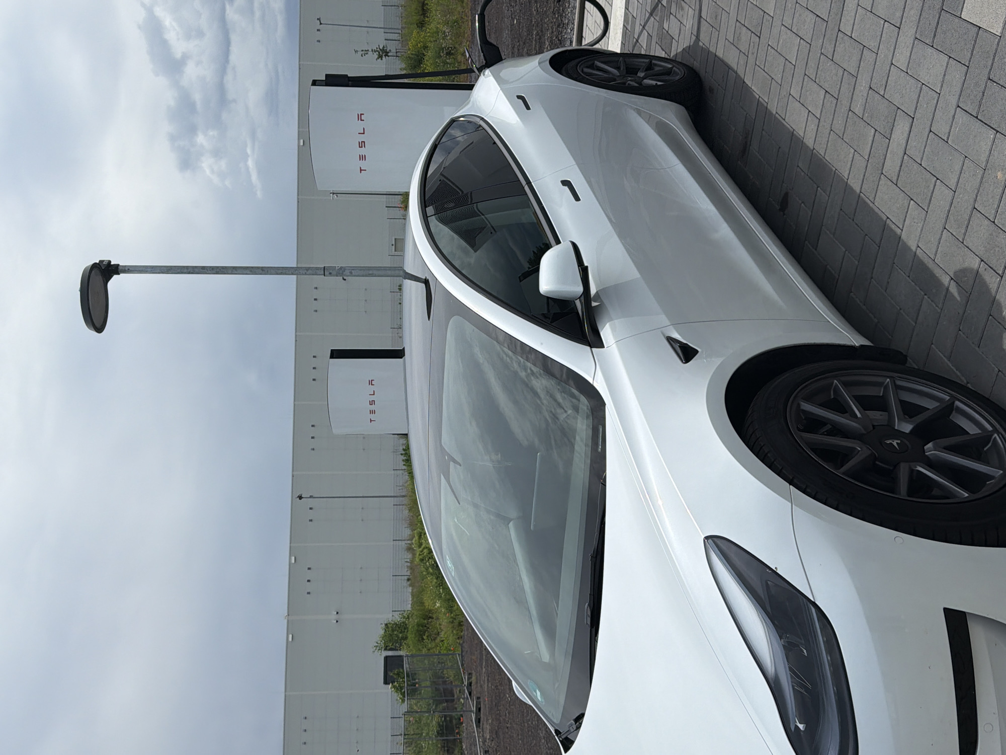The image shows a white Tesla Model 3 parked at a Tesla Supercharger station. The location is Koblenz, Germany. The Tesla chargers are visible in the background along with a portion of a large building. A lamppost is present on the left side of the image. The car is parked on a brick-paved surface. The sky is overcast.