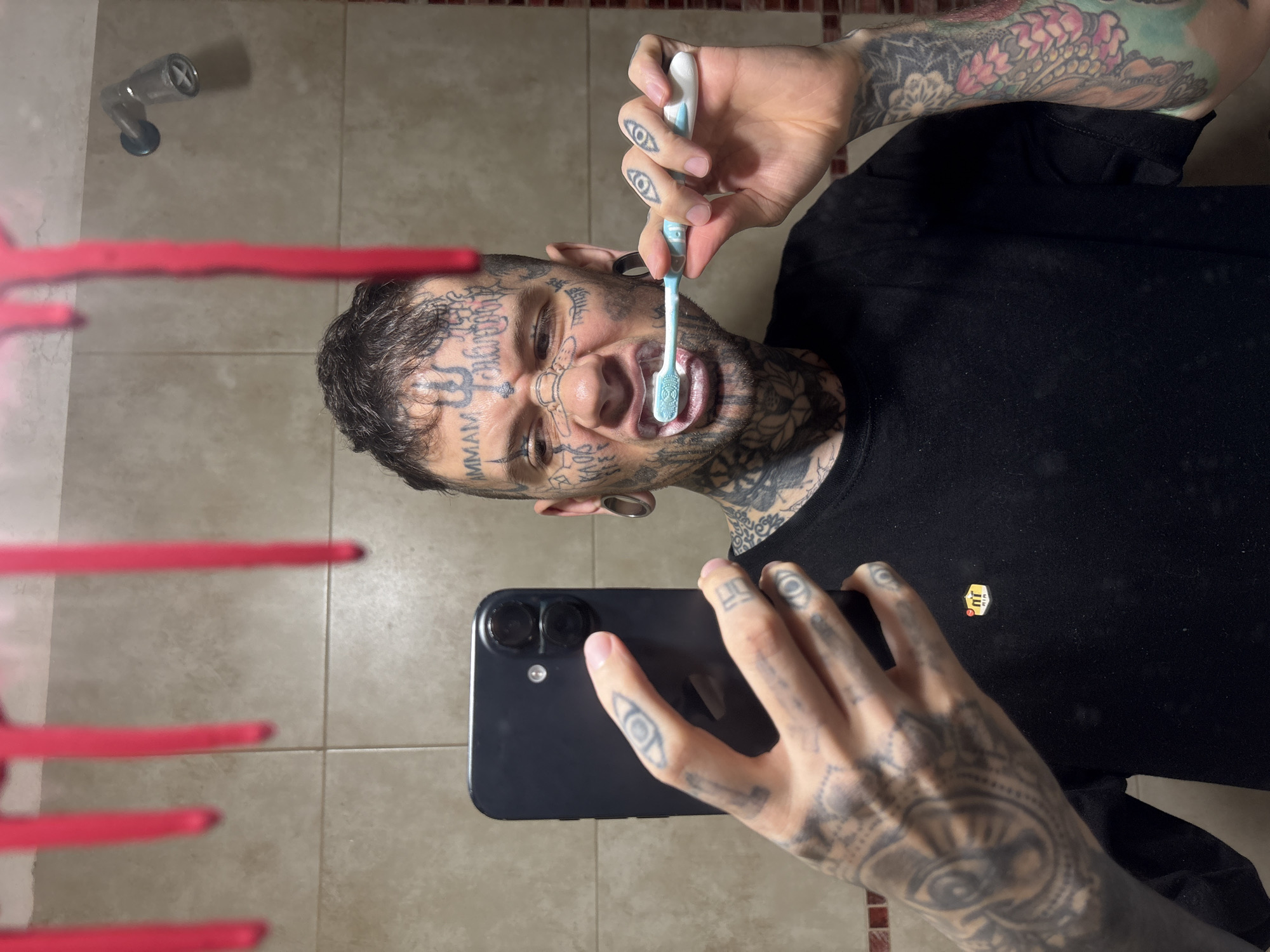 A heavily tattooed male is depicted in a mirror selfie, brushing his teeth within a bathroom. He holds a dark-colored smartphone with dual rear cameras in his left hand to capture the image, while operating a light blue and white toothbrush in his right hand; toothpaste foam is visible around his mouth. The individual has extensive dark tattoos covering his face, neck, hands, and arms, large ear gauges, and a flesh-colored strip across his nasal bridge. He wears a black short-sleeved t-shirt featuring a 'TN AIR' logo on the left chest. The background consists of light beige square wall tiles and a horizontal stripe of reddish-brown accent tiles. A chrome showerhead is visible on the upper right. Red, vertical, drip-like marks are present on the mirror's surface in the foreground. No specific visual cues within the image identify a location in Buenos Aires, Argentina.