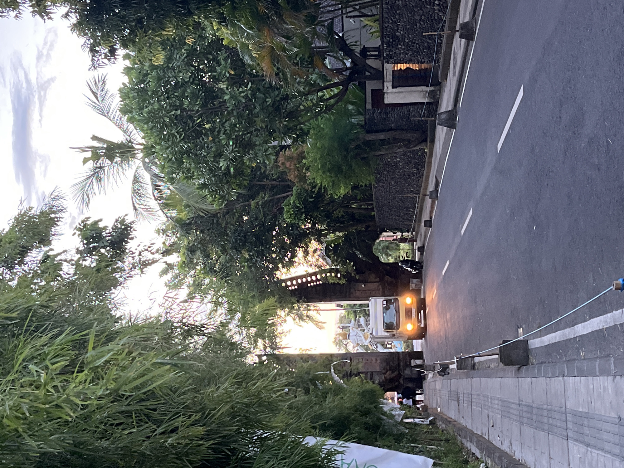The image presents a street scene in Unknown, Indonesia, where a two-lane road stretches into the distance, flanked by a sidewalk on the right. A white truck with its headlights on is traveling along the road. Pedestrians are visible on the sidewalk near a decorated stone structure, possibly an entrance or archway. Lush greenery, including palm trees and other foliage, lines both sides of the road. A building with a stone wall is also visible on the right side of the road. The sky is overcast.