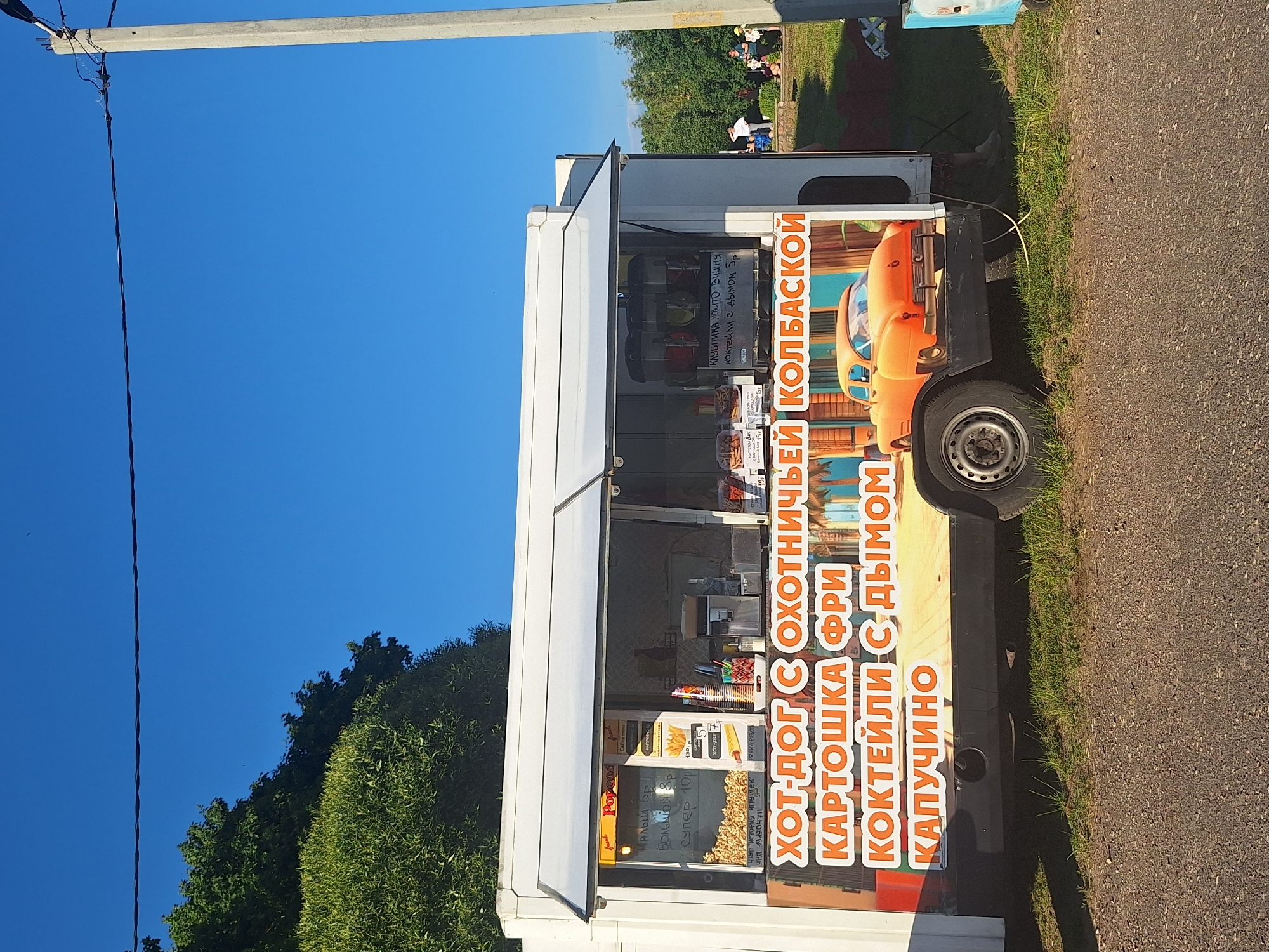 A food truck is parked on a grassy area under a bright blue sky with some trees in the background. The food truck is white with a large graphic on its side featuring an orange classic car and text in Russian. The text advertises various food items like hot dogs, french fries, cocktails, and cappuccino. Inside the truck, there are visible signs with prices and product names. People are seen in the background, some sitting on the grass and others standing, suggesting a public outdoor setting, likely in the vicinity of Цімкавіцкі сельскі Савет, Belarus. The overall scene appears to be a sunny day with a relaxed atmosphere.