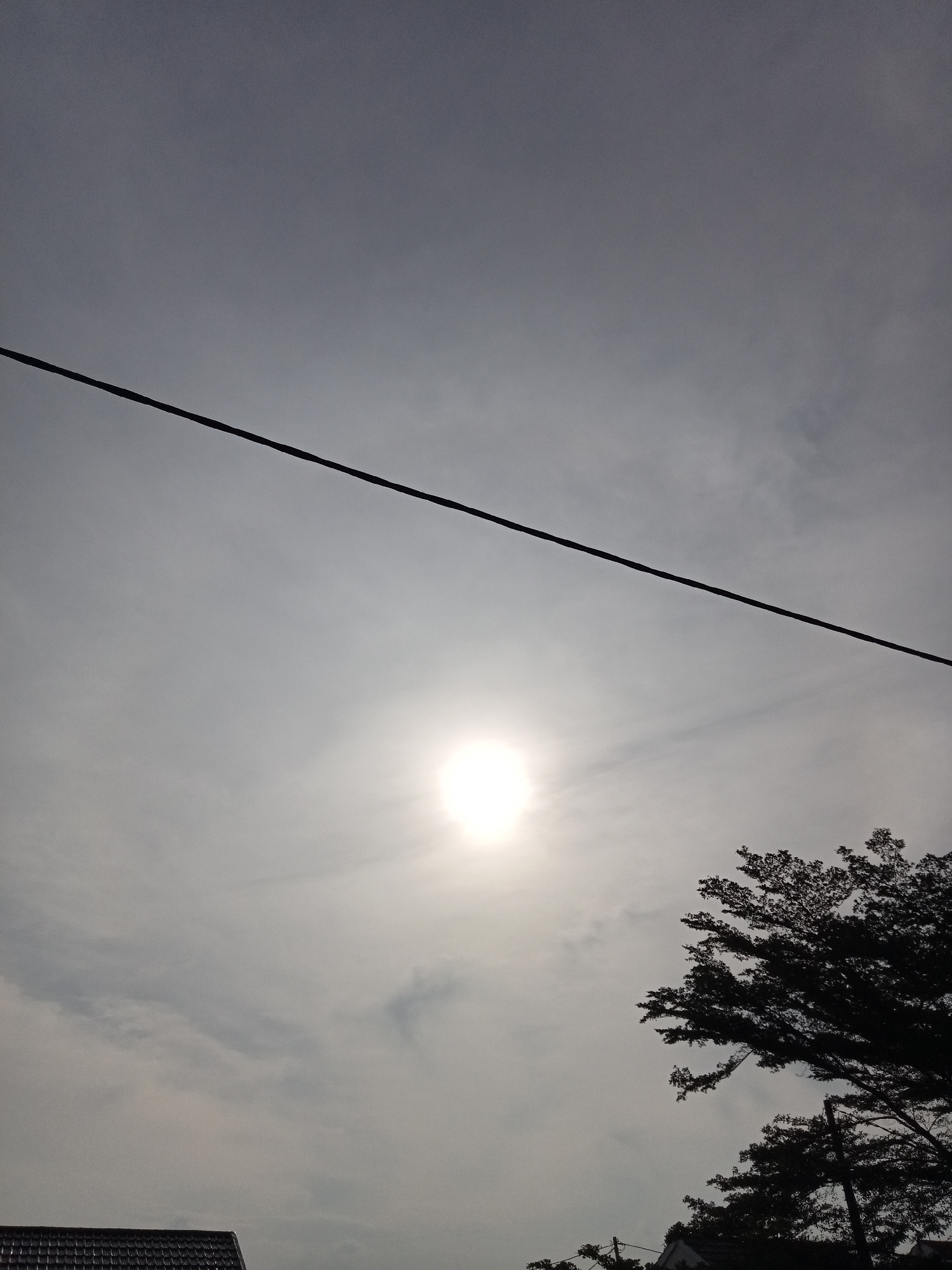 The image captures a bright sun partially obscured by hazy clouds, positioned in the upper center of the frame. A dark, diagonal power line stretches across the upper portion of the image, from the left edge towards the right. In the lower right quadrant, the silhouette of a large tree with dense foliage is visible against the bright sky. The lower left corner shows a sliver of a dark-tiled rooftop, likely belonging to a house. The environment suggests an outdoor scene in the city of Kualu, Indonesia, likely during daylight hours given the sun's prominence. There are no people or discernible actions taking place in the scene.