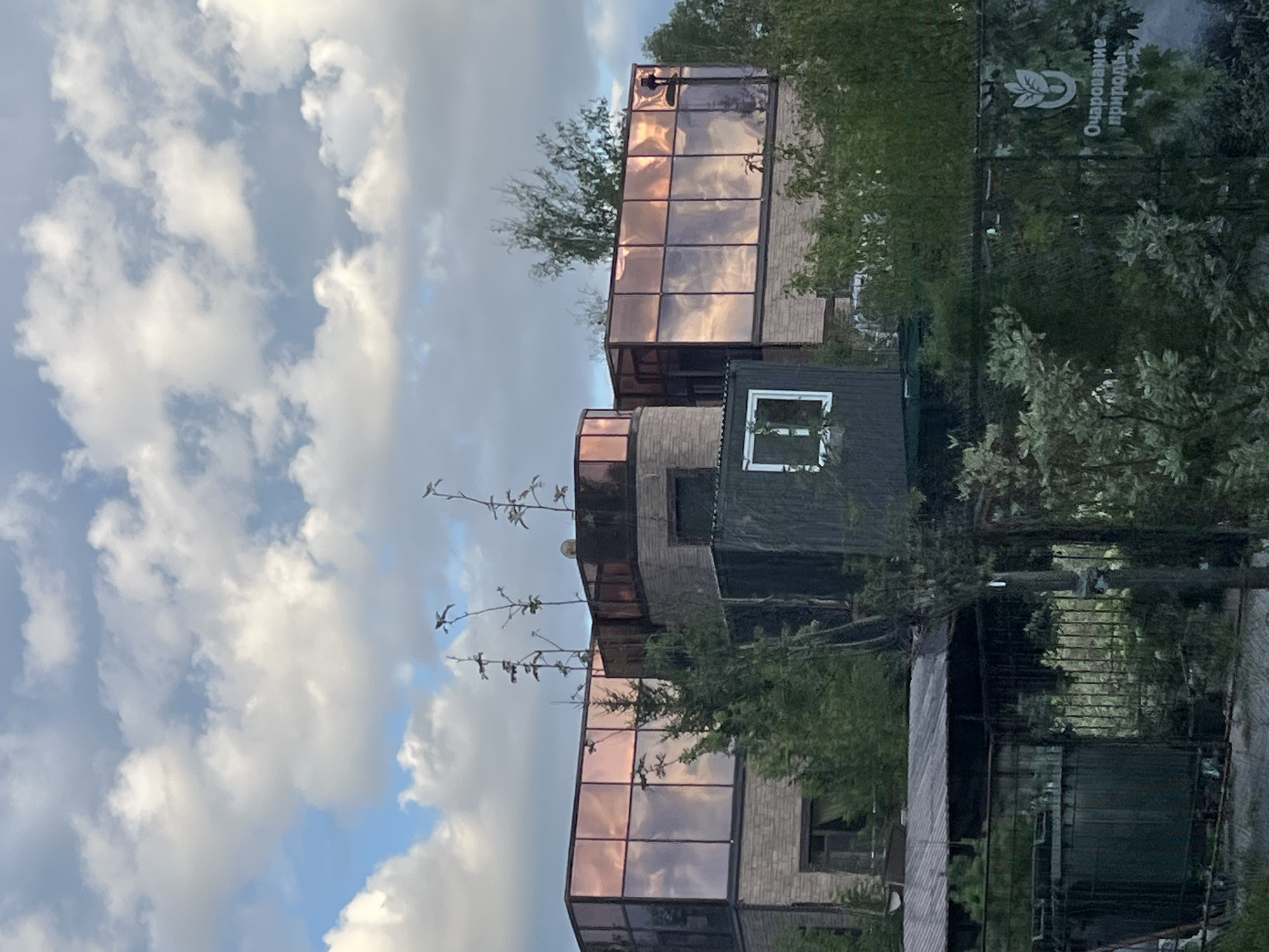 In the city of Ozernyi, Russia, a multi-story building with a modern architectural style is partially visible under a partly cloudy sky. The building features a brick facade and large, reflective windows with a rose-gold tint, which mirror the surrounding clouds. Several thin, bare tree branches extend upwards in front of the building, interspersed with evergreen trees. In the foreground, a dark, rectangular structure with a white-trimmed window is situated in front of the main building. Further down, a fence separates the property from what appears to be a garden or nursery area, with a sign on the fence featuring a leaf emblem and Russian text that reads "Очарование природы" (Charm of nature). A large, dark green bin is visible behind the fence on the left side of the image. The overall scene depicts a building and its surrounding grounds, suggesting a commercial or residential property with an emphasis on natural elements.