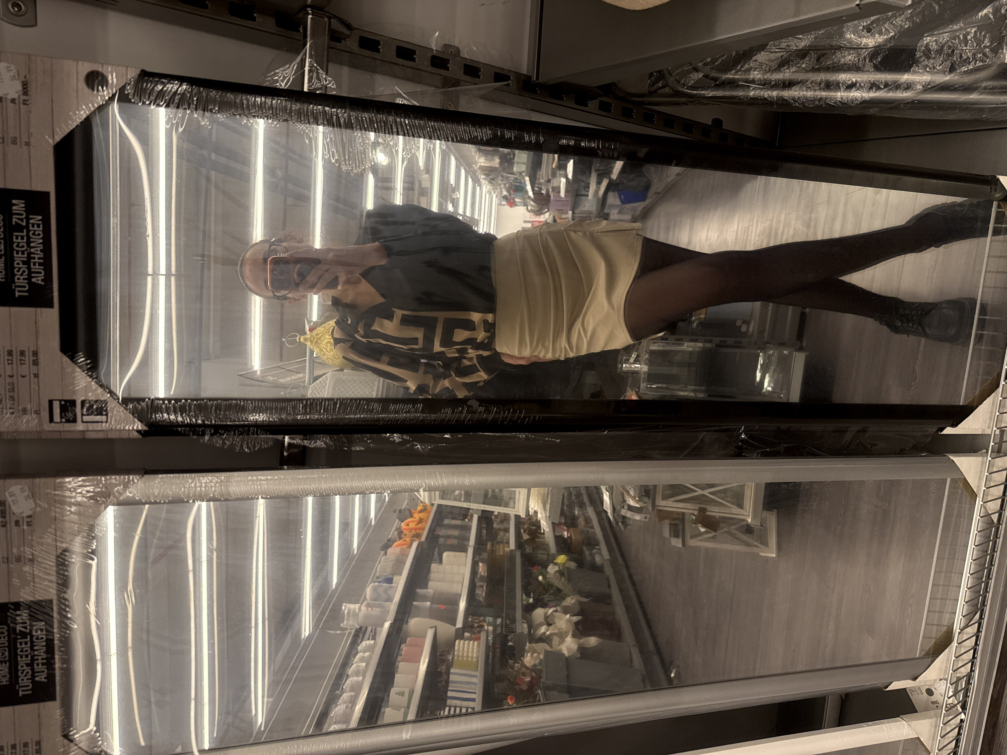 A person is captured taking a selfie reflection in a large, packaged mirror within what appears to be a retail store. The individual, whose face is partially obscured by a smartphone, wears glasses, a dark top with a distinct geometric pattern, a light-colored skirt or skort, dark tights or leggings, and dark boots.
Two large mirrors, still encased in protective plastic wrap, are visible, prominently displayed in the store aisle. Labels on the mirrors clearly state "HOME DECO" and "TÜRSPIEGEL ZUM AUFHÄNGEN," which is German for "DOOR MIRROR FOR HANGING." The reflection in the mirror reveals a long store aisle lined with shelves. These shelves hold various home decor items, including what look like small towels, candles, artificial flowers, and some orange pumpkin-shaped decorations, suggesting seasonal offerings. The store features a light-colored wooden floor and bright overhead fluorescent lighting, typical of a modern retail environment.
Visible text on the mirror packaging includes multiple price points and currency indicators: "€ 17.99," "34.99," "Ft 8000-," and "KC. 495.00." The abbreviation "BG" is also present, which could signify Bulgaria, aligning with the provided location context of Pleven. Another text "AFISK SLO" suggests a potential connection to Slovenia.