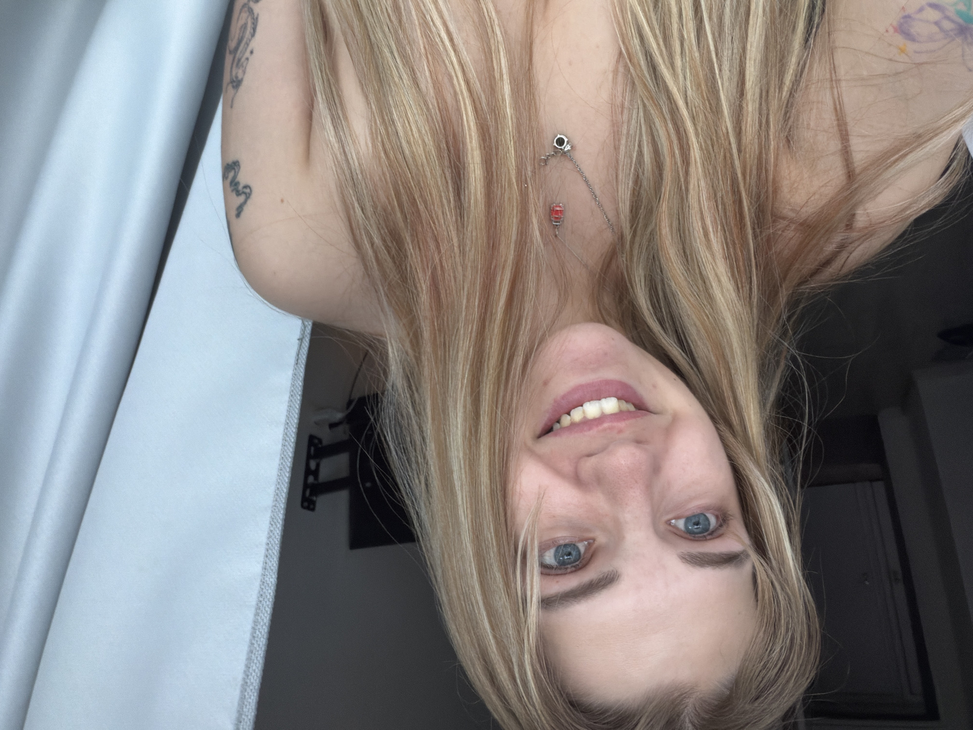 A close-up, inverted selfie features a fair-skinned person with light blonde hair, blue eyes, and visible tattoos on their upper arm and shoulder. A necklace with a pendant is visible. The background is comprised of a white curtain and a dark interior space. The location in the city of Kazan, Russia is not determinable from this image.