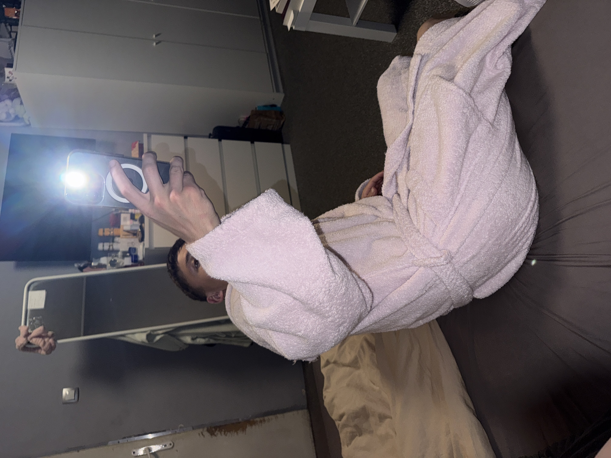 A person wearing a light pink terrycloth robe is reclining on a bed with grey sheets. The person is holding a smartphone, with the camera flash activated, pointed towards a television in the background. The room features white cabinets and shelves with various items on them. A beige colored duvet is partially visible near the person on the bed. The location of Sofia, Bulgaria is not discernible from the image.
