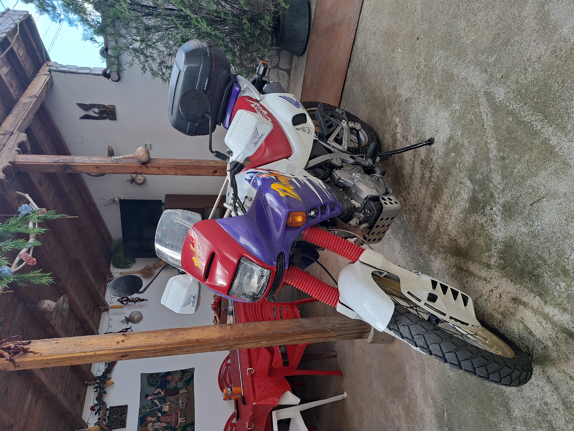 A Honda motorcycle stands parked on a concrete surface in an outdoor setting, likely a patio or courtyard, in the city of Unknown, Unknown. The motorcycle has a red, white, and purple color scheme with the Honda logo prominently displayed. It features a top case mounted on the back. The surrounding area includes a covered patio with wooden beams and support posts. Decorations such as gourds and other objects are attached to the wall behind the patio. Red plastic chairs and tables are partially visible in the background. The setting suggests a residential or commercial outdoor space.