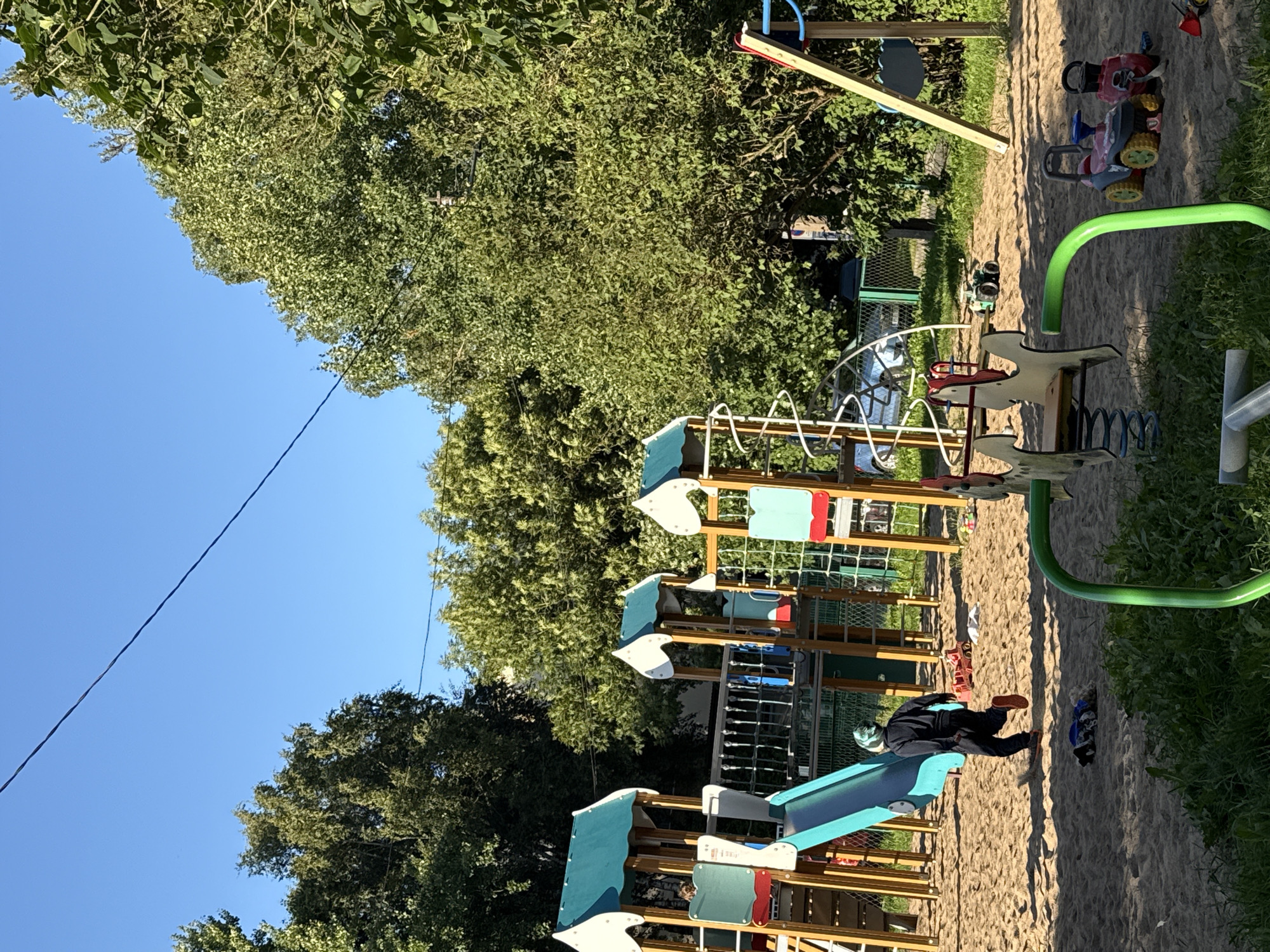 In the city of Unknown, Unknown, on a sunny day, a child in a dark tracksuit and a blue and green patterned hat runs across a sand-covered playground. The child is heading away from a multi-level play structure with a blue slide, green and white roofs, and ladders. Scattered in the sand are various toys, including a red toy truck, a green toy vehicle, and two ride-on toys – one pink and blue, and one red. A green metal spring rider for children is visible in the foreground, partially obstructing the view of the sand. Beyond the playground equipment, lush green trees form a dense canopy under a clear blue sky. A thin wire stretches diagonally across the sky.