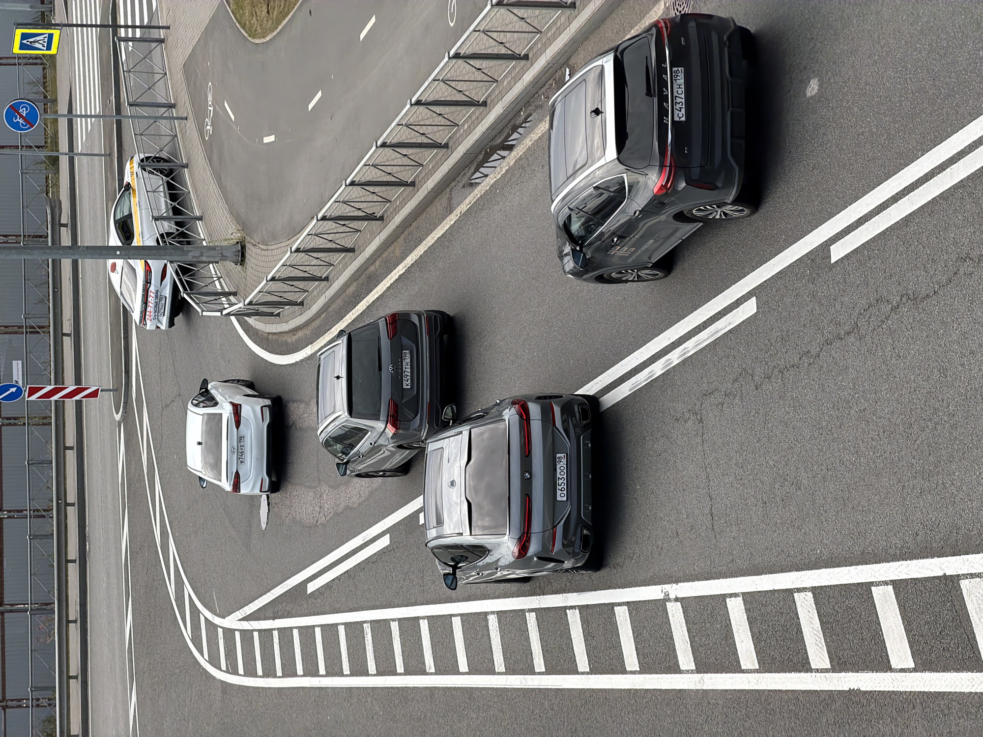 The image depicts an overhead view of a road intersection or roundabout in Saint Petersburg, Russia, likely in an urban or suburban area given the presence of multiple lanes, traffic markings, and safety barriers. A white sedan with a yellow stripe and taxi markings is turning right. Behind it, a dark gray SUV is traveling straight, followed by another dark gray SUV. In the foreground, a dark gray coupe-style SUV is also moving straight. The road surface is asphalt, marked with white lines indicating lanes and lane divisions. Metal barriers line the sides of the road, and a pedestrian crossing with a marked crosswalk is visible in the background, along with traffic signs. The lighting suggests it is daytime.