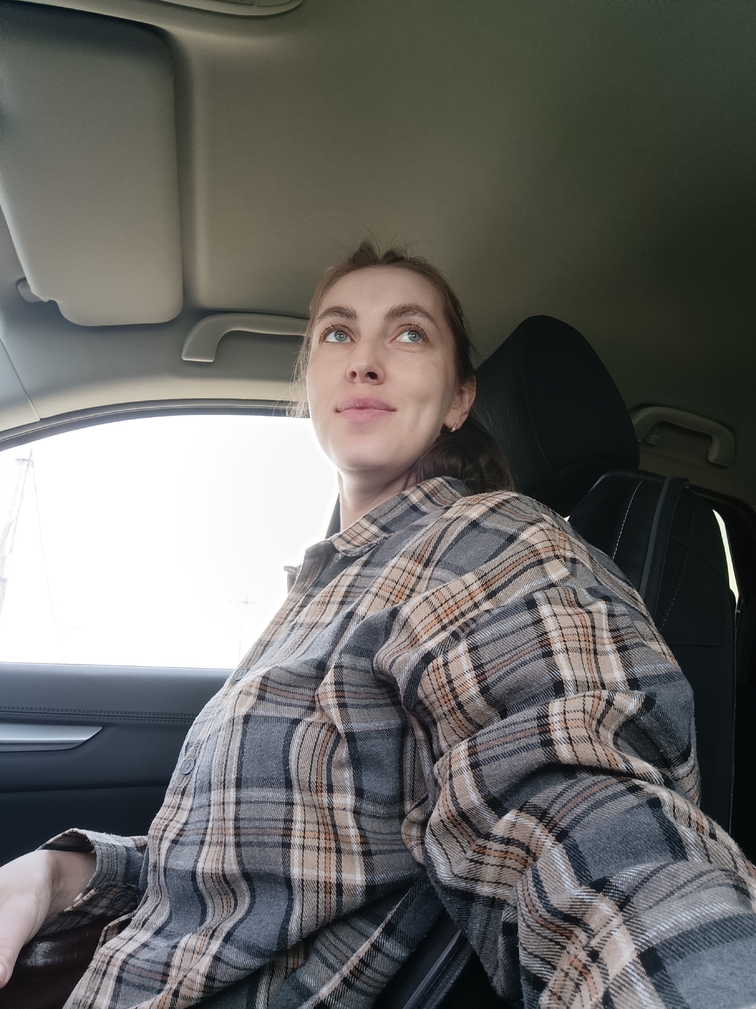 A woman sits in the driver's seat of a car. She wears a gray and tan plaid flannel shirt and is looking upwards toward the car's ceiling and windshield. The car is parked, and the background outside the window appears bright. The interior of the car is dark. The location is not specified in the image, and it is not possible to determine from the image alone that the location is Chaikovsky, Russia.