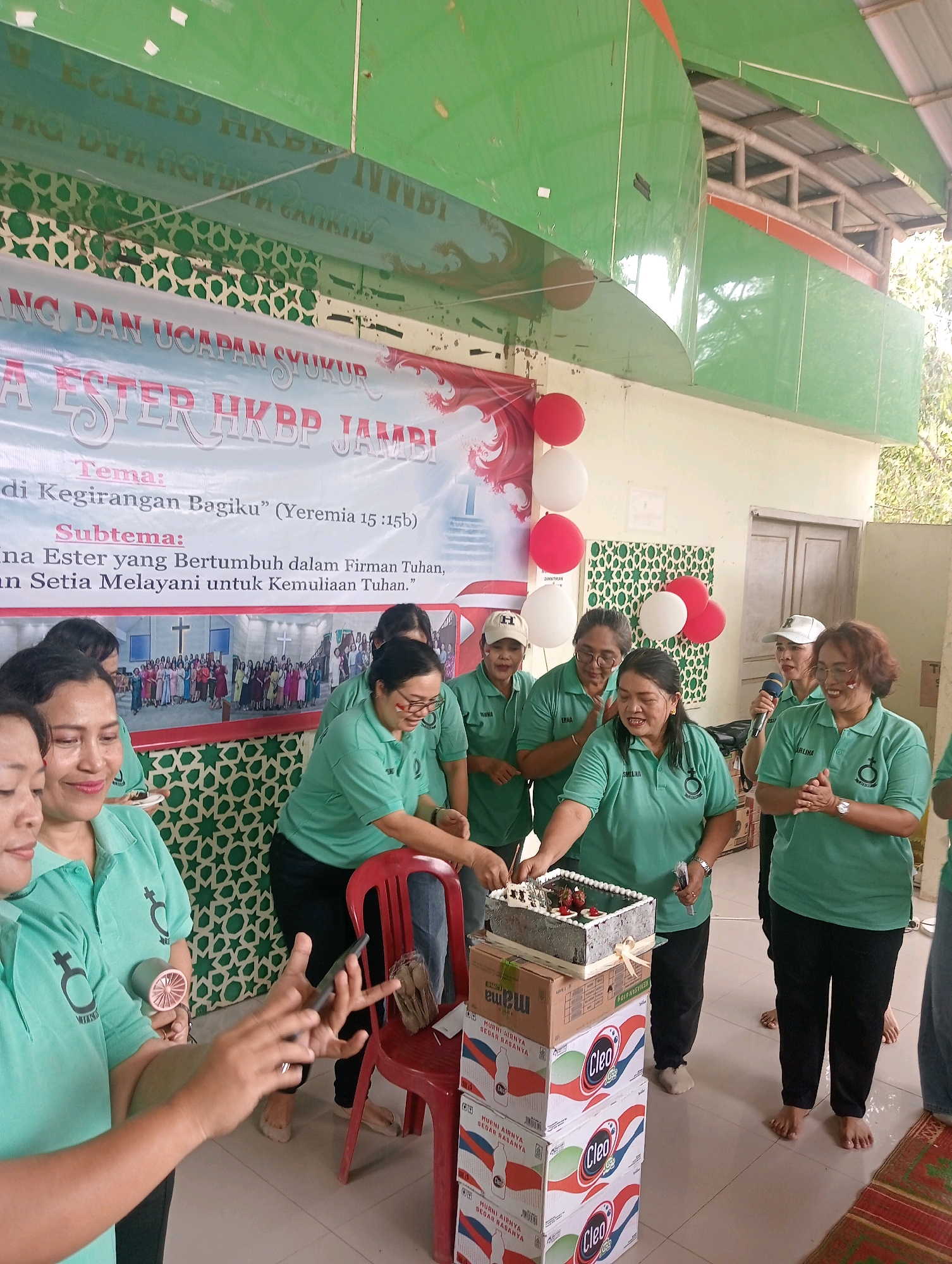 The image depicts an outdoor event with a group of women in matching green shirts, some of whom are wearing glasses and hats. The women are gathered around a cake and stacked boxes, suggesting a celebration or a gathering. In the background, a banner reads "ESTHER HKBP JAMBI," indicating the event is related to the HKBP church in Jambi, Indonesia. Red and white balloons are visible, adding to the festive atmosphere. One woman is holding a microphone, implying a speech or announcement is taking place. The environment appears to be an open area with a roof overhead, possibly a community hall or an outdoor gathering space. The overall scene suggests a joyous occasion involving members of the Esther congregation of HKBP Jambi.