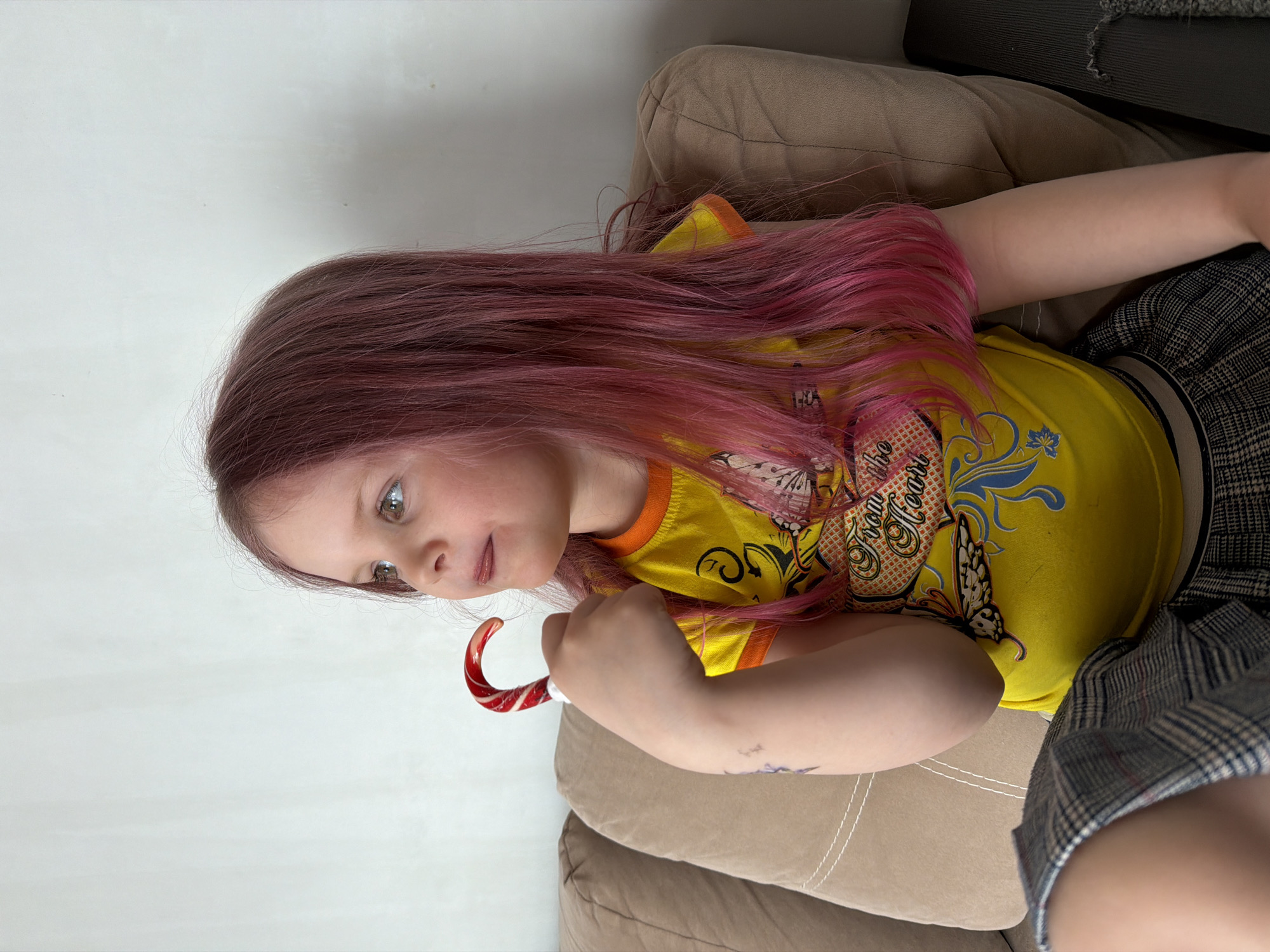 A young girl with pink hair is sitting on a beige couch, holding a candy cane in her left hand. She is wearing a yellow t-shirt with butterfly and floral designs and a plaid skirt. The background consists of a white wall and a gray object. The location is Chaikovsky, Russia.