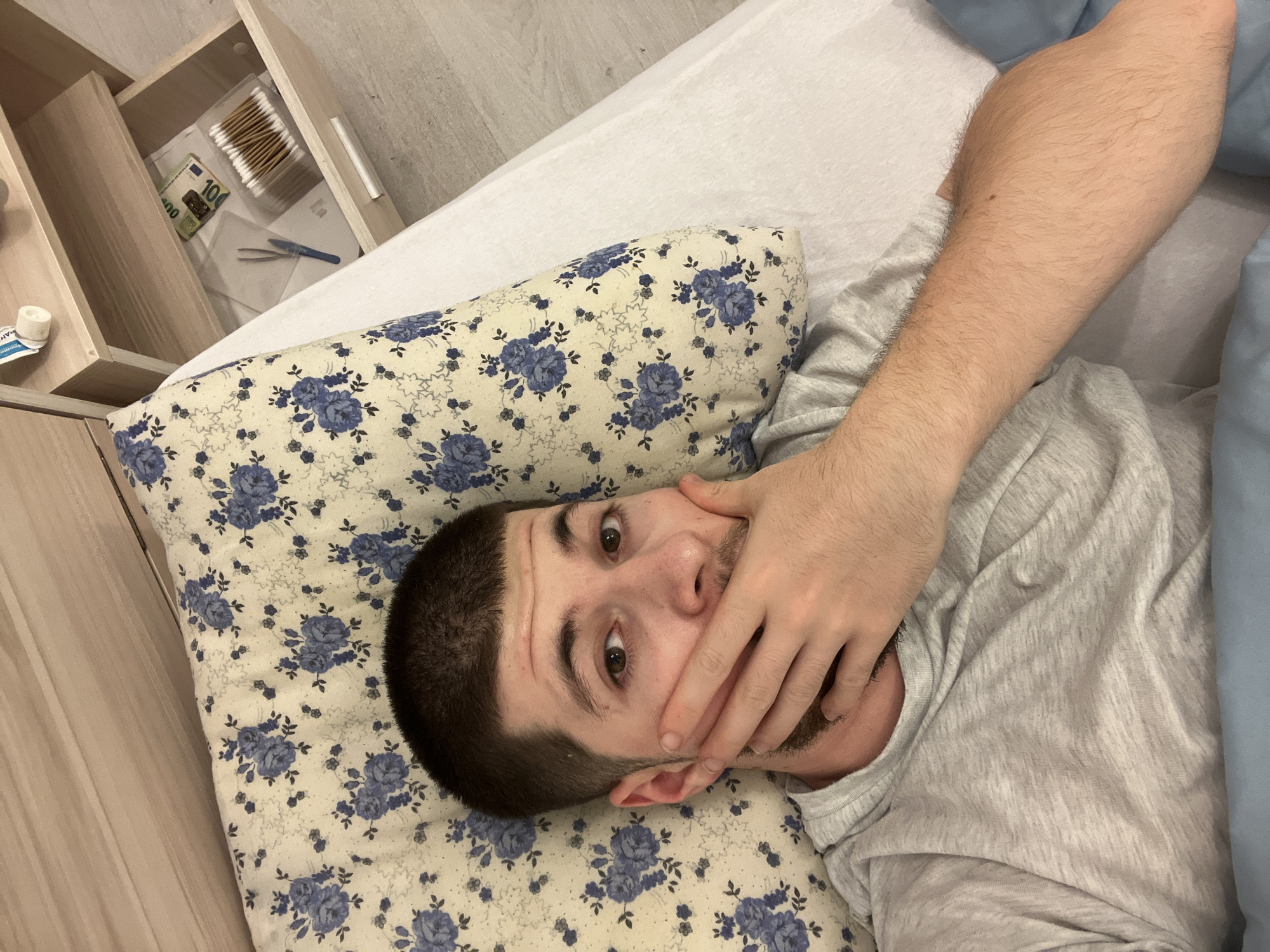 A man with a short, dark haircut lies on a bed. He wears a gray t-shirt and his face is partially obscured by his right hand. His head rests on a pillow with a floral pattern of blue flowers. Beside the bed, a partially open drawer reveals a box of cotton swabs, a pair of tweezers, and a 100 Euro banknote. The location is Varna, Bulgaria, but the image does not provide specific details within the city.