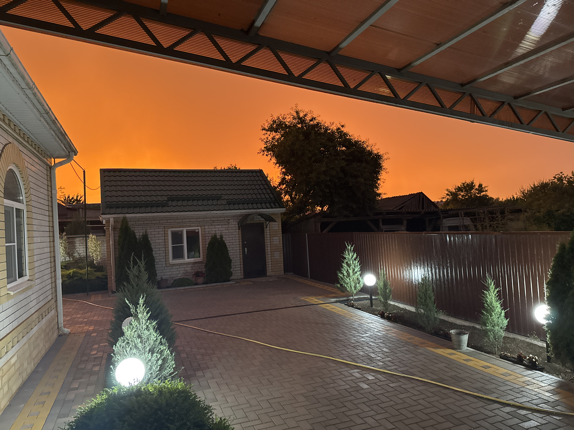 This is an outdoor medium shot of a residential property in Novoselitskoye, Russia, at dusk. The sky is a vibrant orange, suggesting an unusual atmospheric condition like smoke from wildfires or a significant dust storm.
In the foreground, a paved driveway made of brick pavers leads towards a small, single-story building with a dark-tiled roof. A white garage or outbuilding is partially visible on the left. The driveway is lined with small, manicured evergreen trees and modern globe-shaped lights, some of which are illuminated, casting a warm glow on the pavers and surrounding bushes. A yellow garden hose is visible running across the driveway.
Behind the driveway, a dark brown corrugated metal fence runs along the property line, separating it from neighboring areas. Trees and other foliage are visible beyond the fence, silhouetted against the brightly colored sky. A large, leafy tree is prominent in the middle ground. The building directly facing the driveway has a dark door and a window, with more greenery and what appears to be a small potted plant beside it. The lighting in the scene is a mix of ambient twilight and the artificial lights from the property's fixtures. The overall mood is somewhat dramatic due to the intense sky color, creating an unusual and striking visual. There is no visible text in the image.